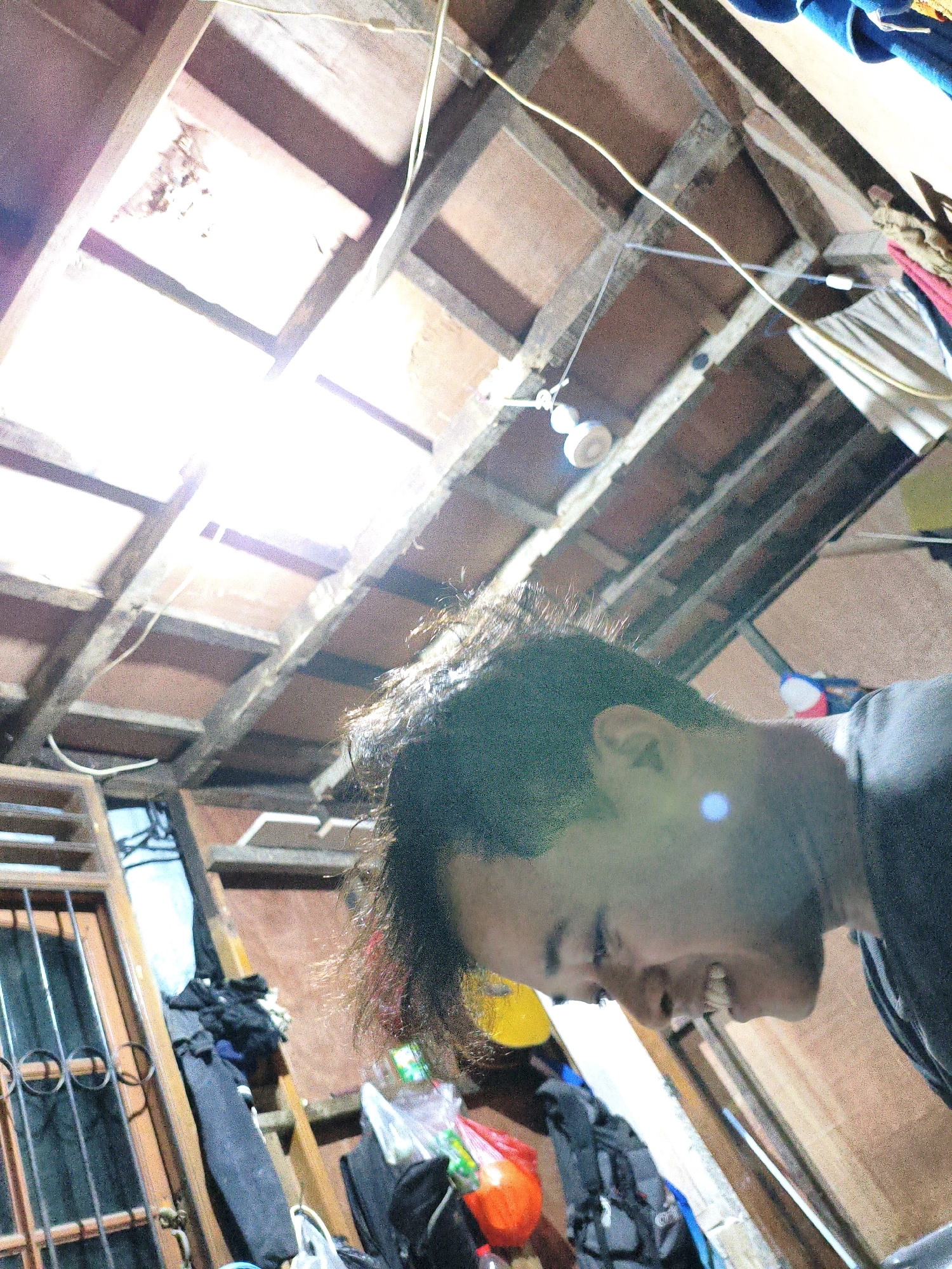 The image depicts an indoor scene with a person's head and shoulders visible from the right side, looking downwards and to the left. The background shows a cluttered, unfinished ceiling constructed of wooden beams and planks, with exposed electrical wires and a bare light bulb. To the left of the person, a wooden structure with metal bars, possibly a window or door, is visible, along with piles of clothes and bags. The overall environment appears to be a dimly lit, possibly residential, space in an urban setting. The presence of unfinished construction elements suggests a utilitarian or perhaps developing area. This scene is identified as being located in the city of Bekasi, Indonesia.