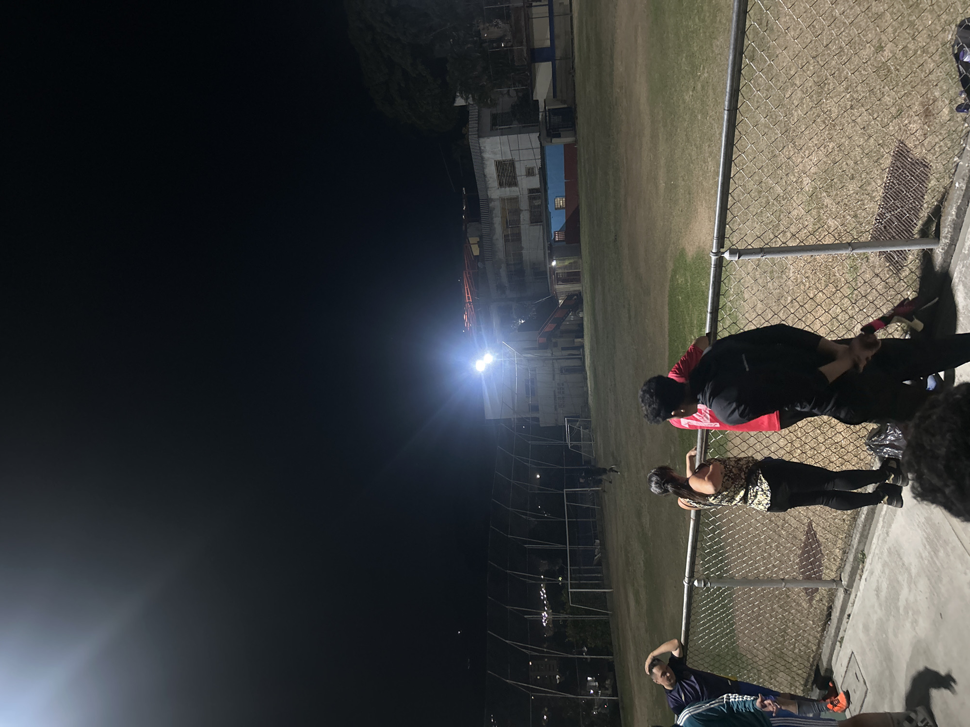 The image captures an outdoor scene at night, likely a sports field in Caracas, Venezuela. The left side of the image is dominated by darkness, while the right side is illuminated by artificial lights.
A chain-link fence runs diagonally across the frame, separating the viewer from the field. Several people are standing near the fence, appearing to be watching or interacting with activity on the field.
Behind the field, there are buildings and trees silhouetted against the night sky. A bright light source illuminates the field and casts shadows. A soccer goal is visible in the distance. The overall scene suggests a casual evening gathering at a sports facility.