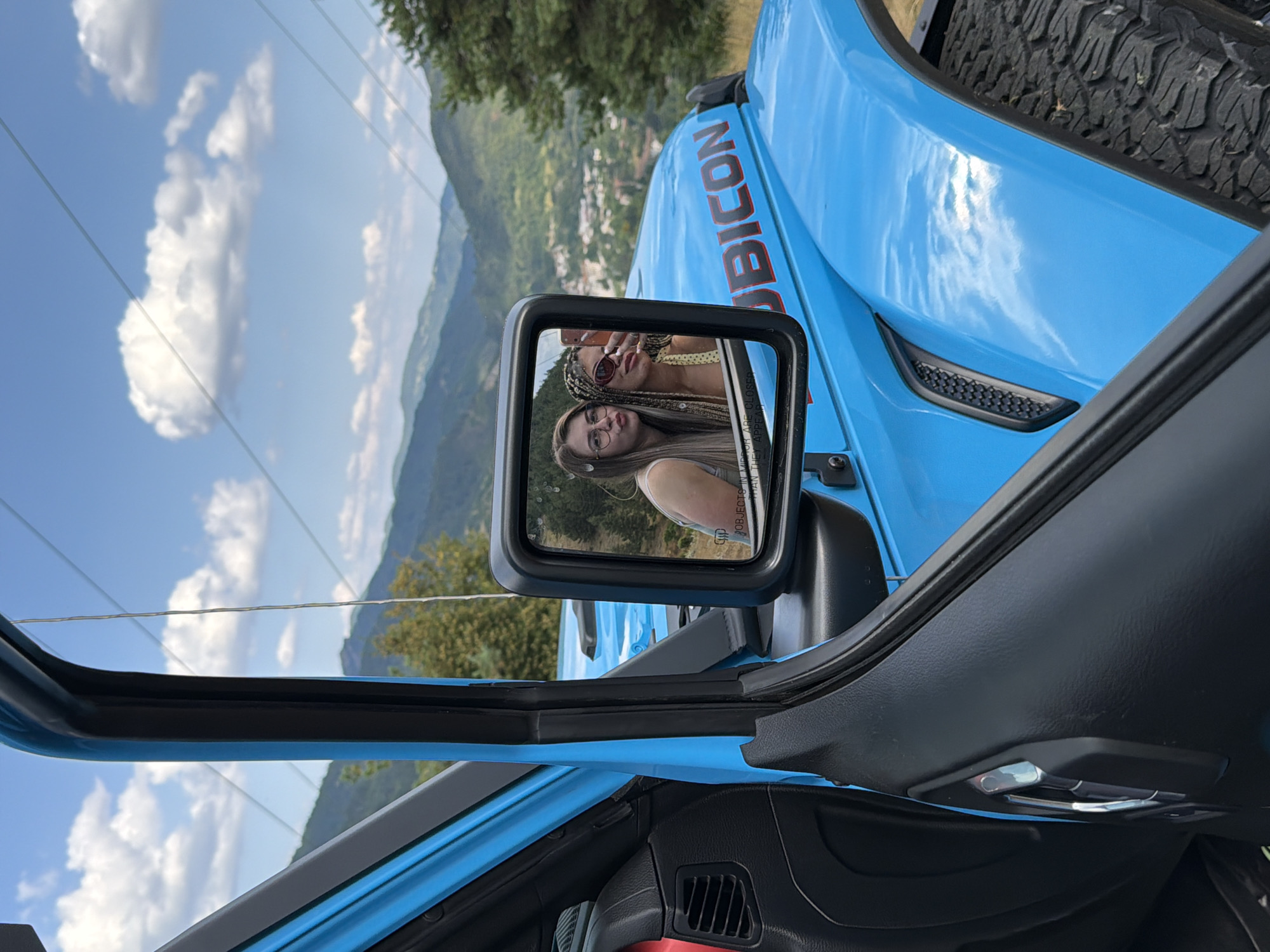 The image captures a bright blue Jeep Wrangler, seen from the driver's side, with the door ajar. The side mirror reflects two young women taking a selfie. The woman on the left has blonde hair and glasses, while the woman on the right has braided hair and sunglasses. They appear to be posing with puckered lips. The Jeep's "RUBICON" decal is visible on the fender.
In the background, a scenic mountain landscape unfolds, with layers of rolling hills covered in lush green trees. A town with white buildings is nestled in the valley below the mountains. The sky is a vibrant blue, dotted with fluffy white clouds. The overall impression is that of a scenic drive or outing in a mountainous region, possibly near Smolyan, Bulgaria, given the terrain and architectural style of the town visible in the reflection. The foreground is dominated by the car's interior and exterior, with the tire and fender of the Jeep clearly visible. The image is shot on a sunny day, with the bright sky and colorful Jeep suggesting a pleasant outdoor environment.