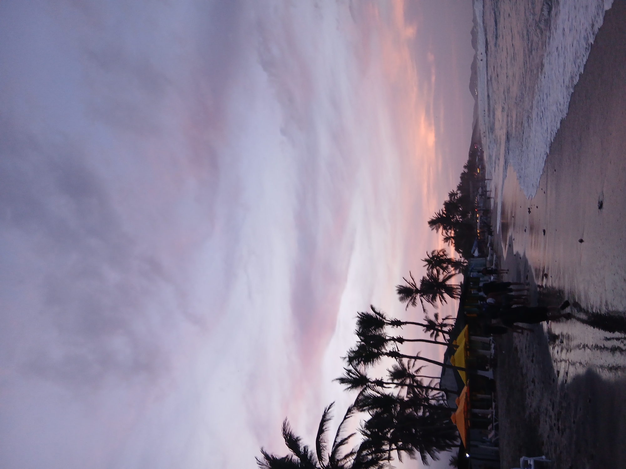 The image depicts a beach at Playa Parguito, Venezuela, at dusk. The sky is a mix of purple and orange hues. Palm trees are silhouetted against the sky along the shoreline. Waves are visible on the right side of the image, meeting the sand. People are walking along the beach near beach umbrellas and chairs.