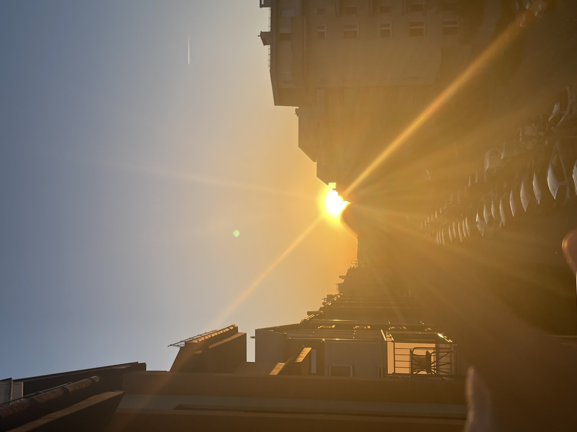 The image depicts a cityscape at sunrise or sunset, with a bright sun positioned between buildings. The sky transitions from blue to orange-yellow as it nears the sun. A thin contrail is visible in the upper left. Buildings of varying heights line both sides of the image, with a row of cars parked on the right. The perspective is from a low angle, looking upwards.