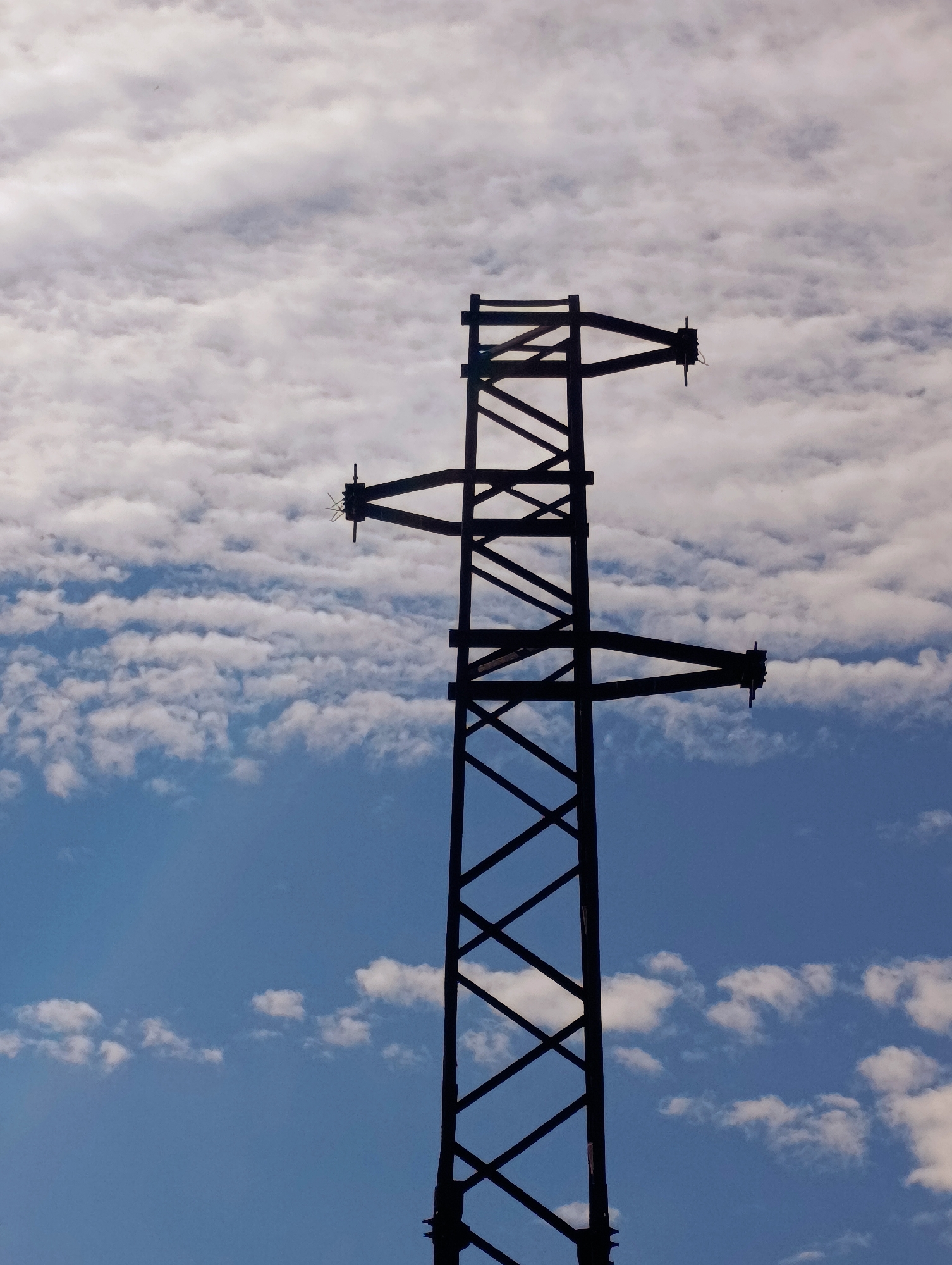 The image shows an electrical transmission tower against a backdrop of a partly cloudy sky, a mixture of blue sky and altocumulus clouds. The tower is silhouetted, revealing its lattice structure and cross arms. The location is possibly Shiroko pole, Bulgaria, based on the prompt. There are no people visible in the image.
