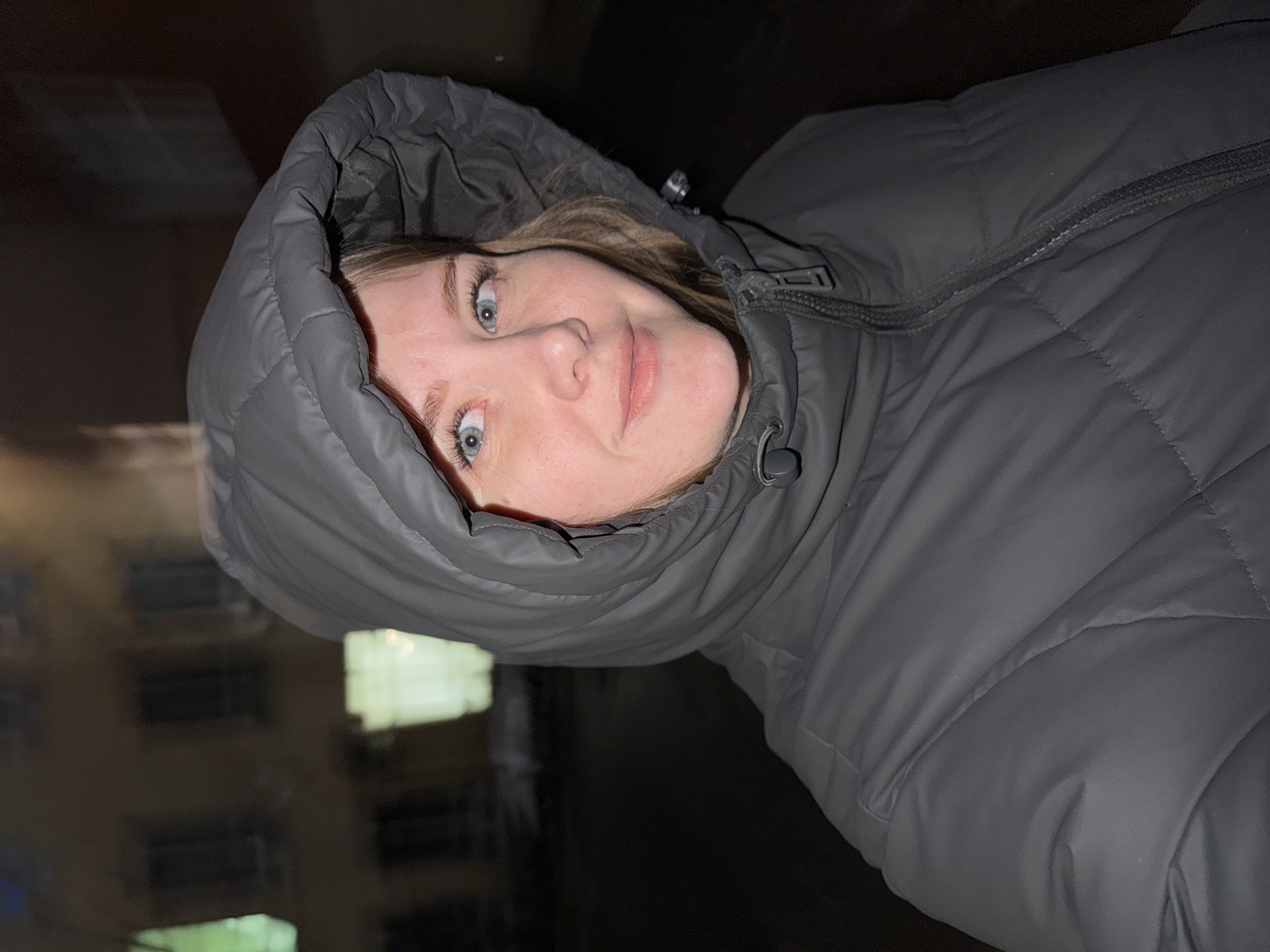 A young woman is depicted in a close-up, portrait-style shot, looking directly at the camera. She has light blue or grey eyes that appear wide, and her expression is a slight, closed-mouth smile or smirk. Her face is framed by the hood of a dark grey, quilted puffer jacket, which she is wearing and zipped up. Her light-colored hair is visible peeking out from under the hood.
The image was taken outdoors at night, likely in an urban setting such as Perm, Russia. The background is largely dark and out of focus, with blurry hints of buildings or windows and faint rectangular light sources, suggesting artificial illumination against the night sky. The presence of a warm puffer jacket implies cold weather conditions. No discernible text is visible in the image.