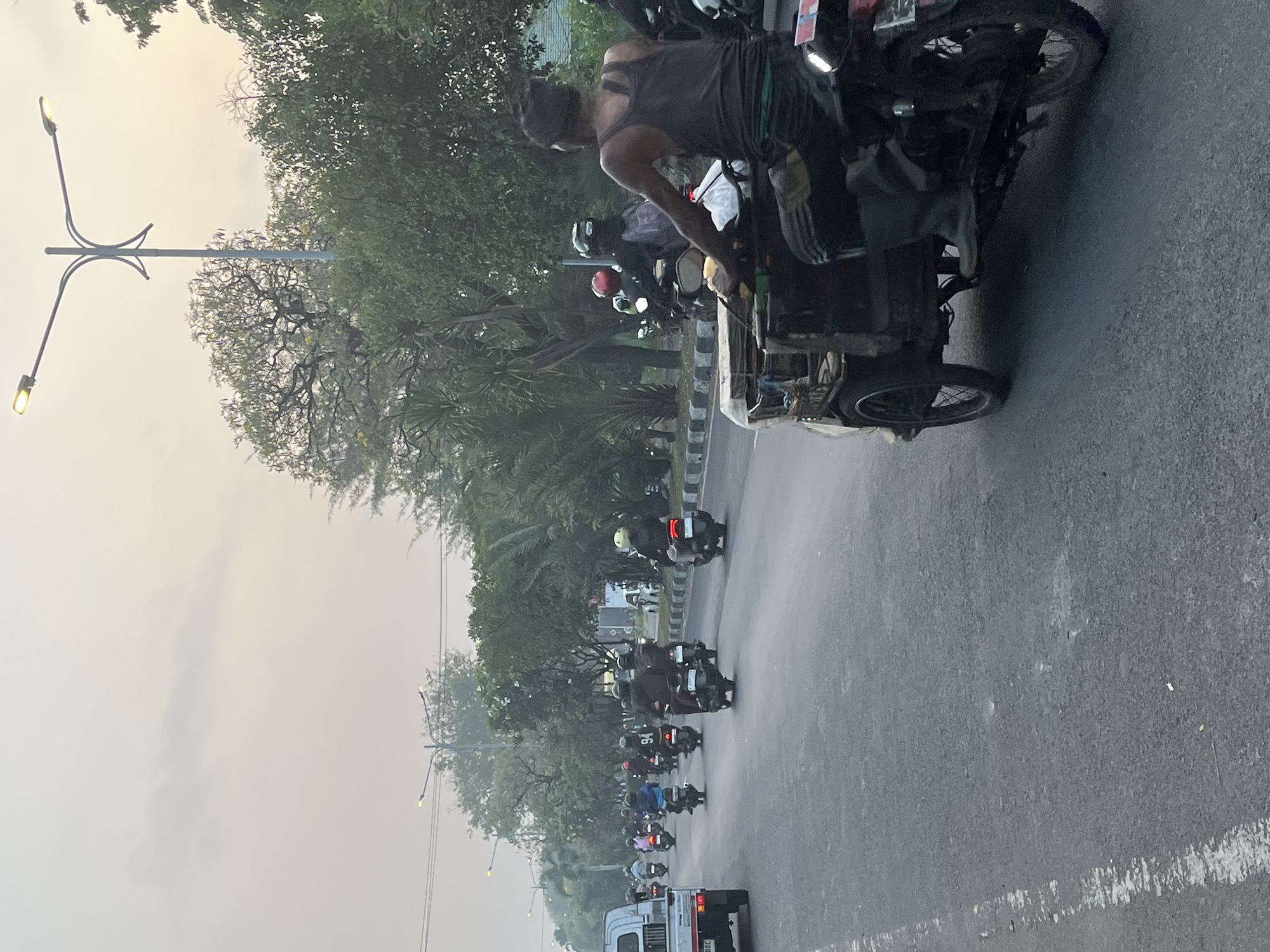 The image depicts a street scene in Kludan, Indonesia, likely during the late afternoon or early morning given the soft, hazy lighting. A procession of motorcycles, many with riders wearing helmets, travels down a multi-lane asphalt road. In the foreground, a three-wheeled motorcycle with a cargo carrier is prominently featured, driven by a man wearing a dark tank top and dark pants with green stripes. He is also wearing dark green boots. Behind him, a line of motorcycles and their riders are visible, some with passengers. The road is lined with trees and palm trees, with streetlights also visible in the background. A white pickup truck is seen on the left side of the road. The overall atmosphere is one of daily commute or movement within the town.