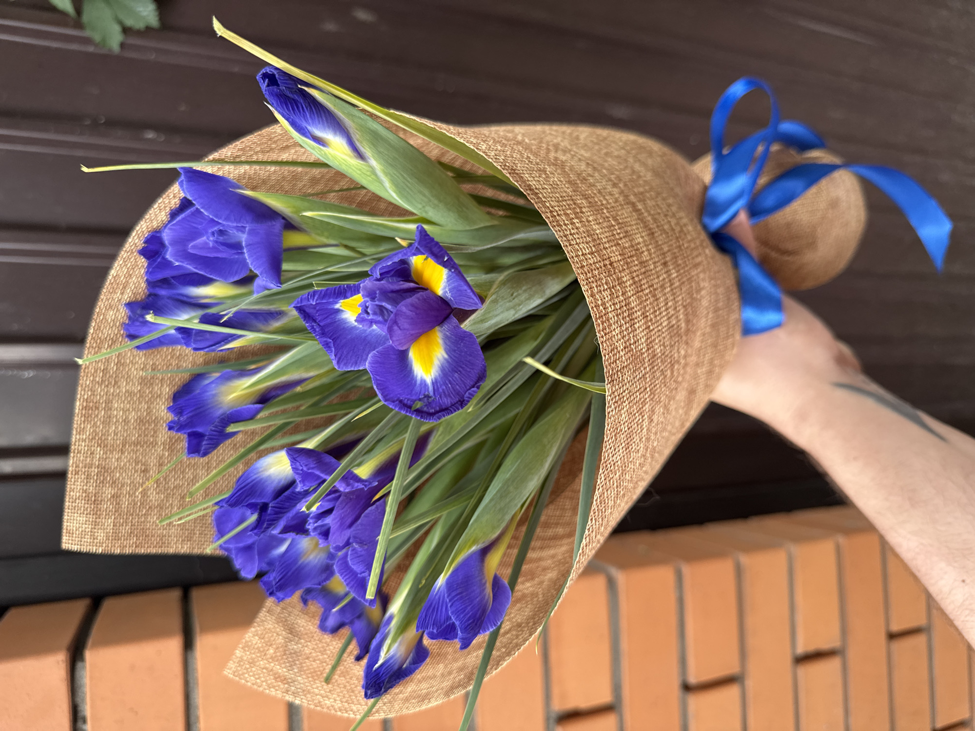 A person's arm and hand hold a bouquet of purple irises wrapped in brown burlap and tied with a blue ribbon. The background shows a brick wall on the left and a dark brown wooden door on the right. The scene appears to be outdoors. The location is not specified in Almaty, Kazakhstan.