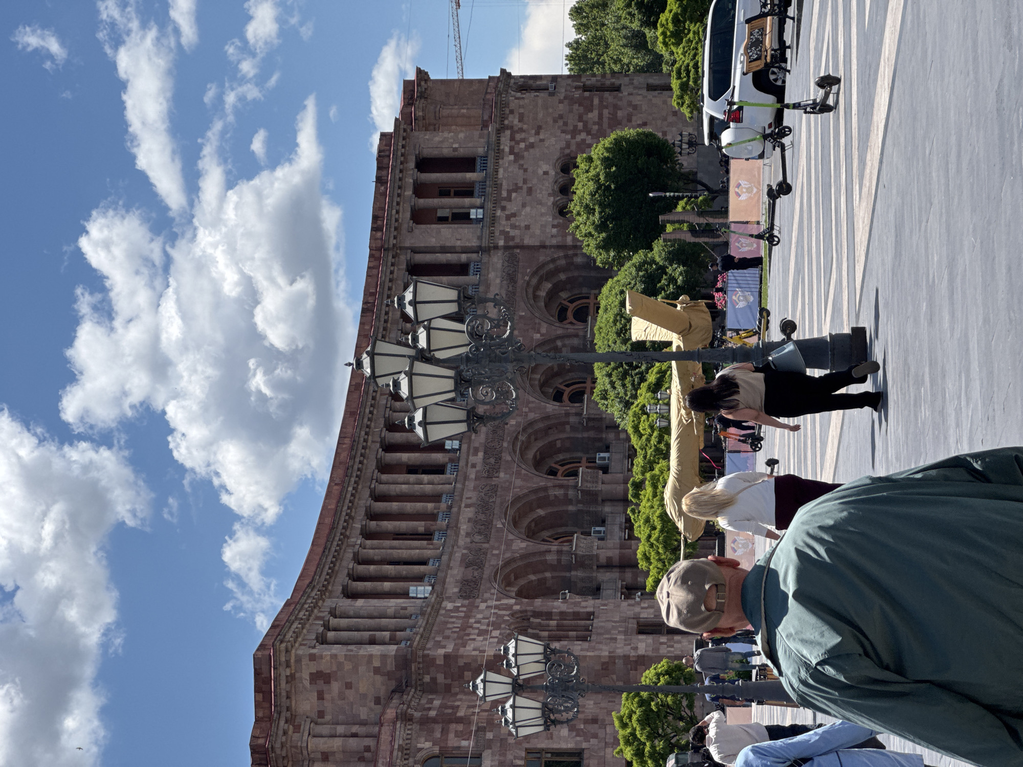 The photograph depicts a street scene in Yerevan, Armenia, featuring a prominent building constructed from reddish-brown stone. The building's facade exhibits arched windows and multiple rows of pillars. A cloudy, blue sky is visible in the background. In the foreground, street lamps are centrally placed. Several people are walking, and there are a few parked electric scooters and a white van visible at the side of the street. The ground is paved with light-colored stones. The environment is urban and well-lit.