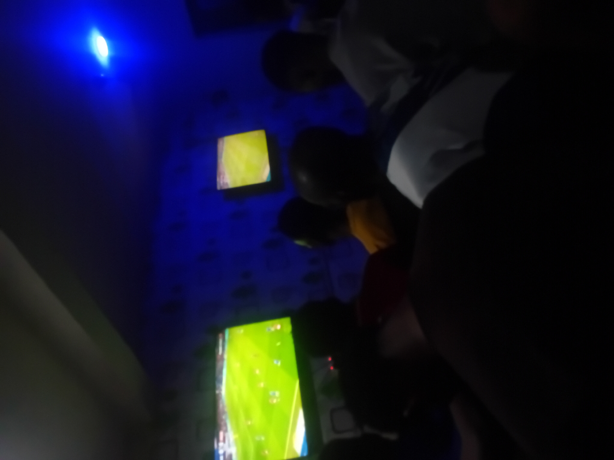 In a dimly lit room with blue-toned walls and a bright blue light source overhead, a group of individuals is gathered, seemingly watching multiple television screens. The screens display what appears to be a football match. The people are partially obscured by shadow, making it difficult to discern details. The overall atmosphere suggests a social gathering focused on watching sports, although the exact location within Lokoja, Nigeria, is indiscernible from the image.