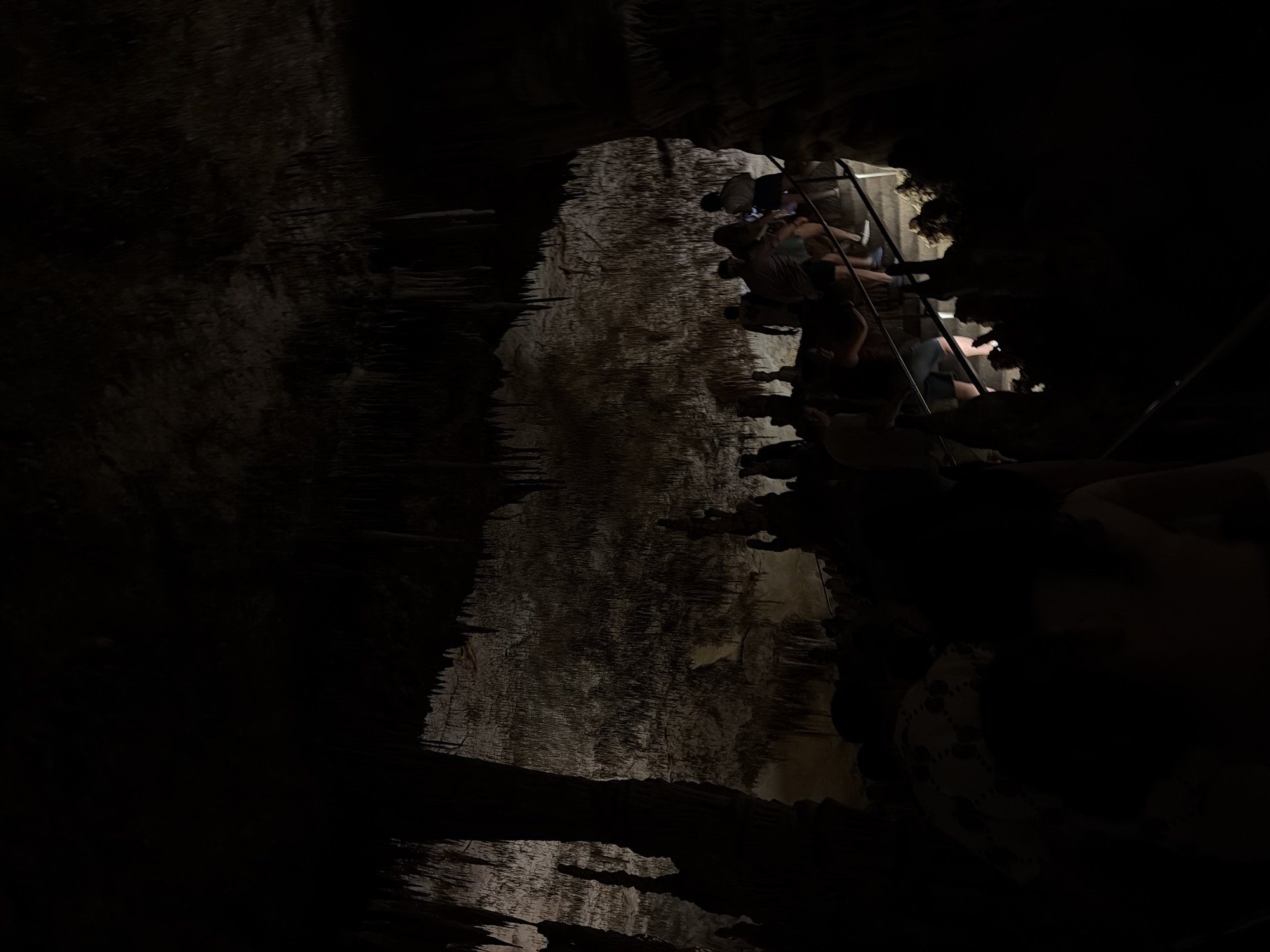 The image shows a view within the Caves of Drach, located in Manacor, Spain. The interior of the cave is dark, with rock formations and stalactites visible in the dimly lit areas. In the upper right of the image, a group of people are climbing a metal ladder.