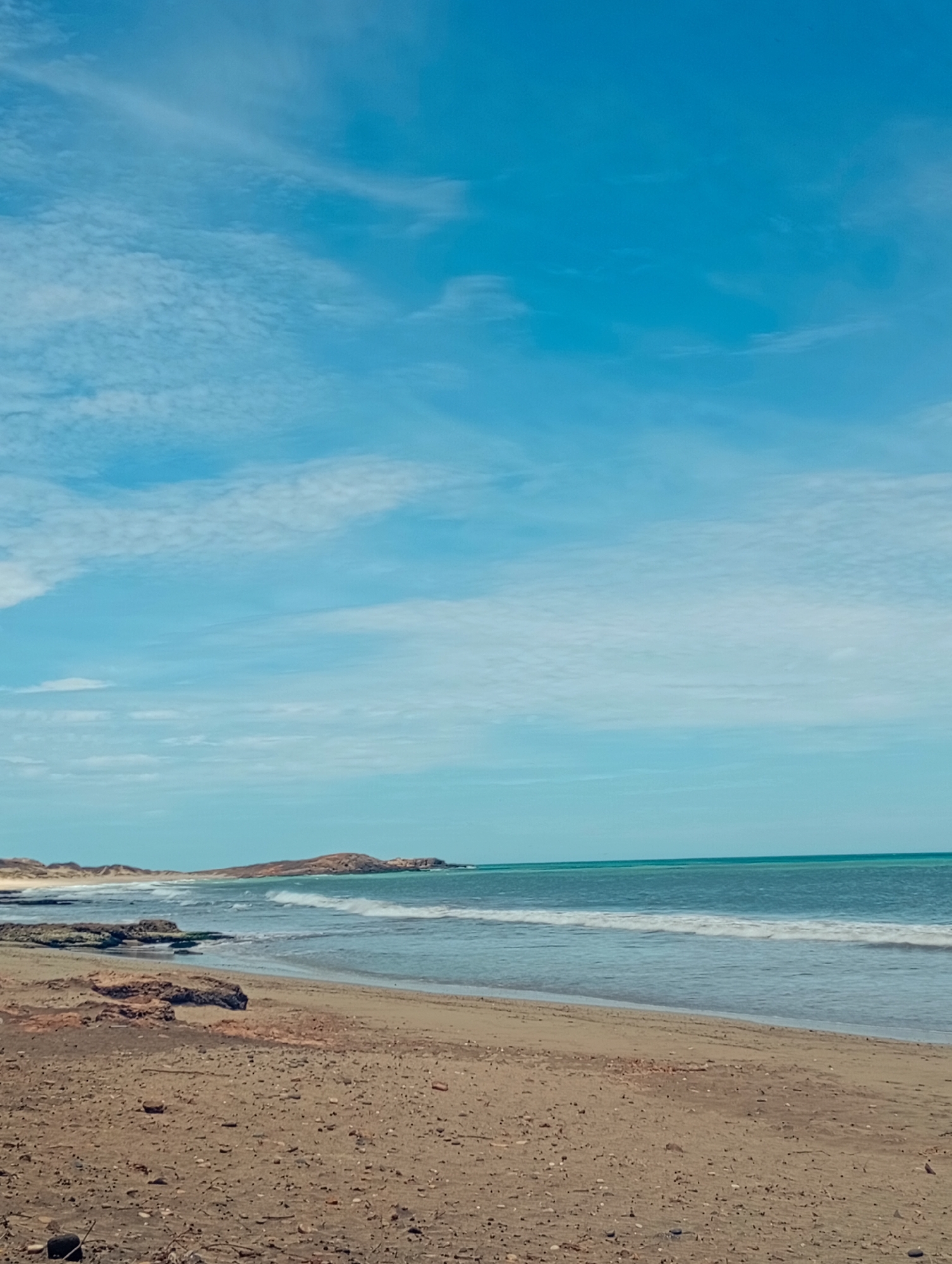The image depicts a coastal scene in Altagracia, Venezuela. The foreground features a sandy beach with small pebbles and rocks. The tide is coming in, and small waves are visible washing onto the shore. In the middle ground, there's a glimpse of a rocky coastline leading to a small headland in the distance. The sky is predominantly blue, with wisps of white clouds scattered across it. The overall tone is peaceful and serene, capturing a typical day at the beach.