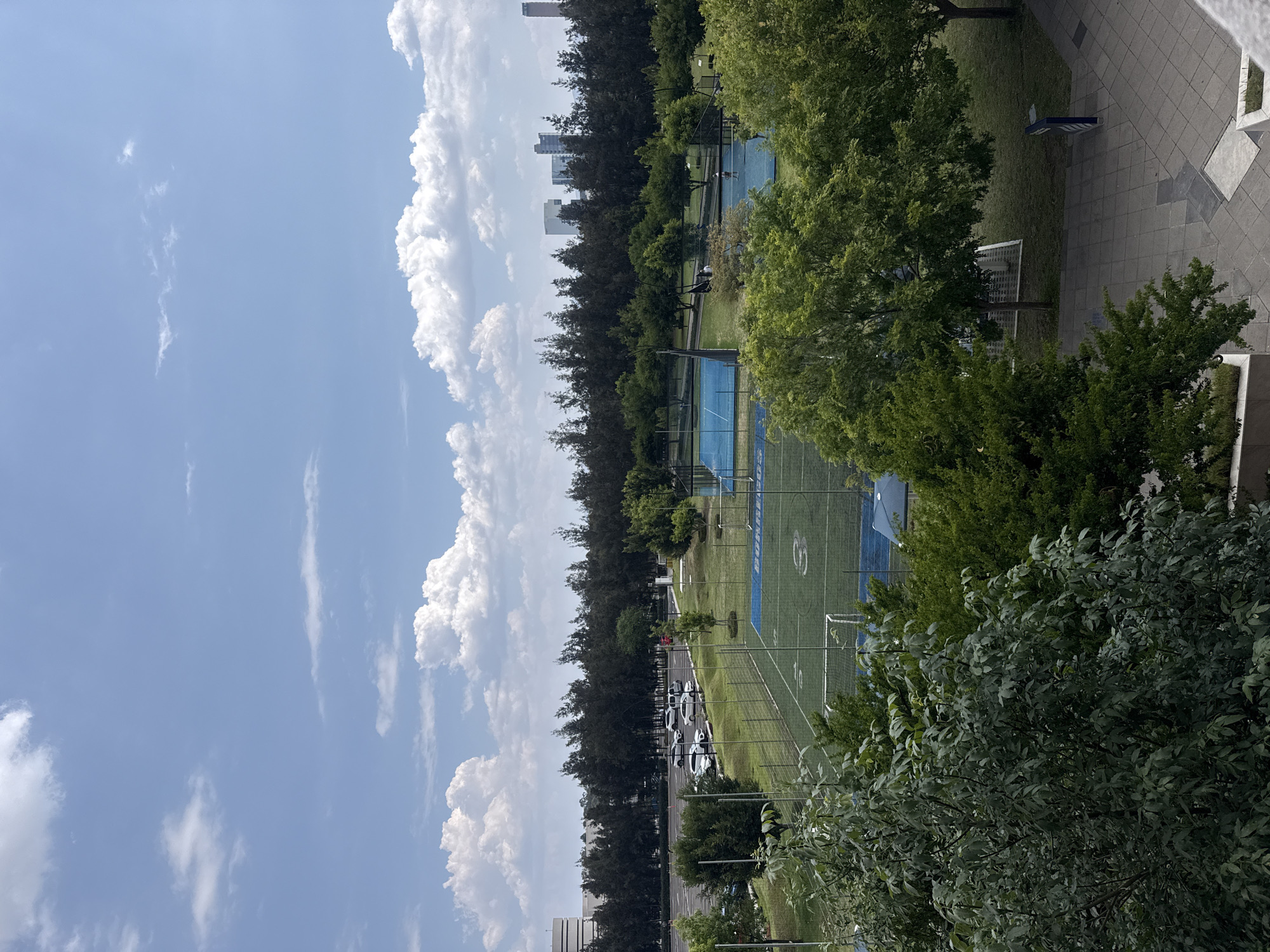 The image captures an elevated, outdoor view in an urban setting. The location is potentially near a city as suggested by distant buildings along the skyline in the background, although the specific city is unknown. A blue sky with fluffy, cumulus clouds provides the backdrop, while trees are on both sides of the scene, framing the field of view. The primary focus is on two sports fields: a rectangular green field is adjacent to a blue-surfaced court. Additionally, a row of parked cars can be seen along the edge of the field, next to the tree line.