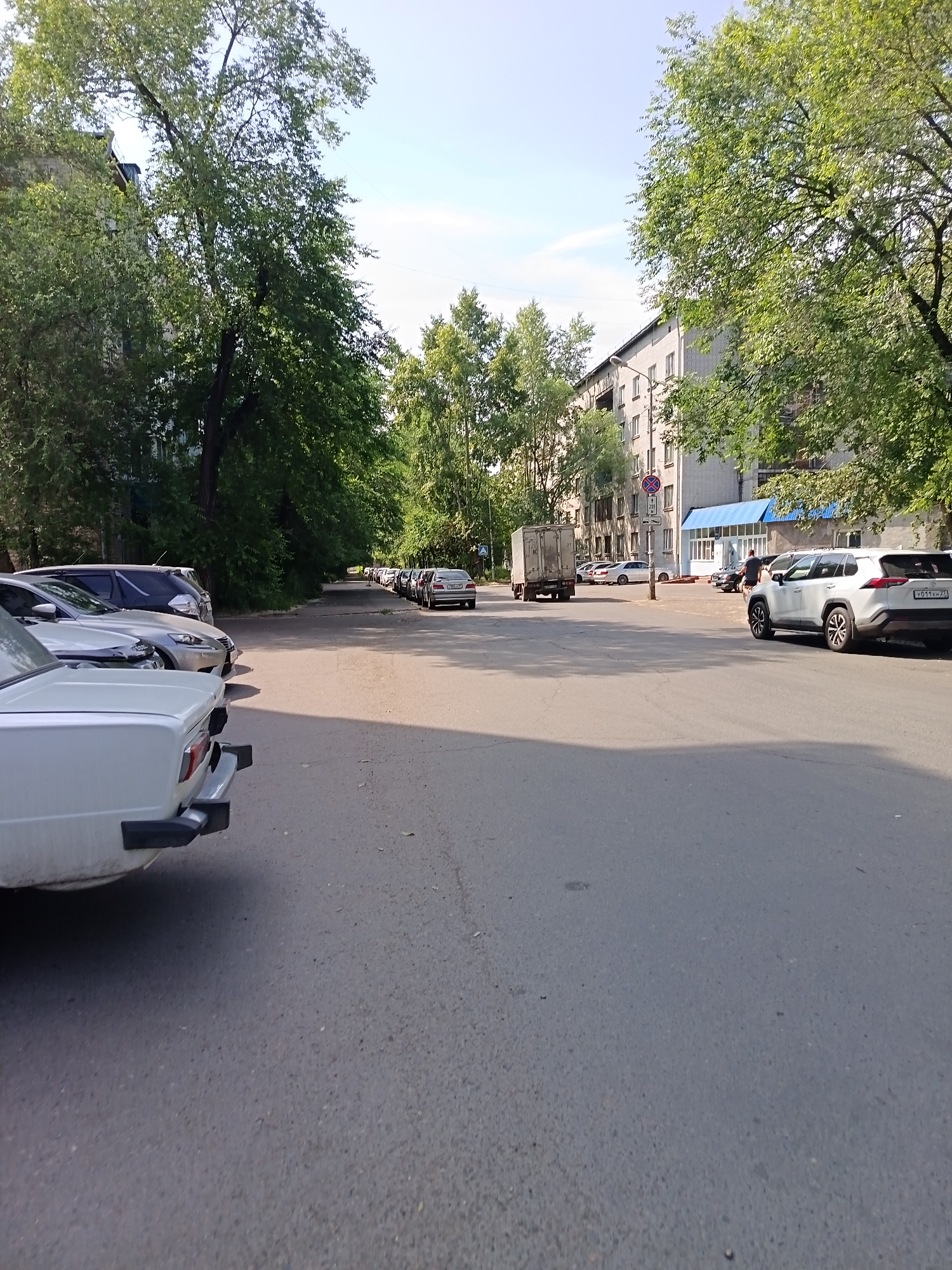 The image shows a street scene in the city of Komsomolsk-on-Amur, Russia. A white car is parked on the left side of the road, with its rear bumper facing the camera. Further down the street, a line of parked cars is visible on the right side of the road. A white truck with a trailer is parked on the left side of the road. A building with a blue awning is visible in the background. The street is paved and lined with trees. The sky is clear and blue.