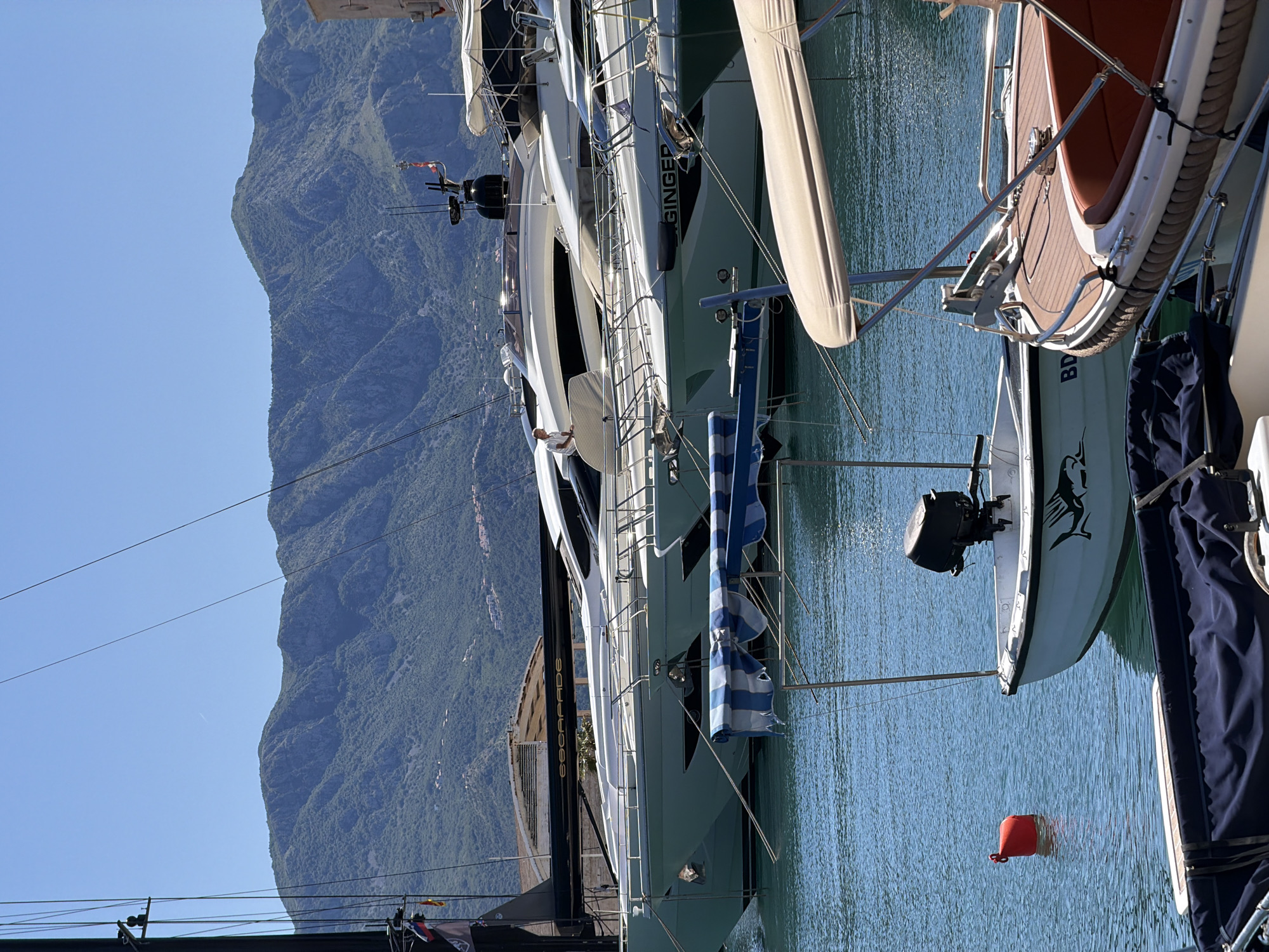 The image captures a scene in Budva, Montenegro, featuring a row of docked yachts in a marina, set against a backdrop of a mountain range and a clear blue sky.
The yachts are of varying sizes and designs, including one labeled "GINGER." Some have awnings or covers, and one has a small dinghy attached to the stern. The water in the marina is calm, reflecting the sunlight.
In the background, the mountain range rises steeply, covered in green vegetation. The sky is cloudless and blue.