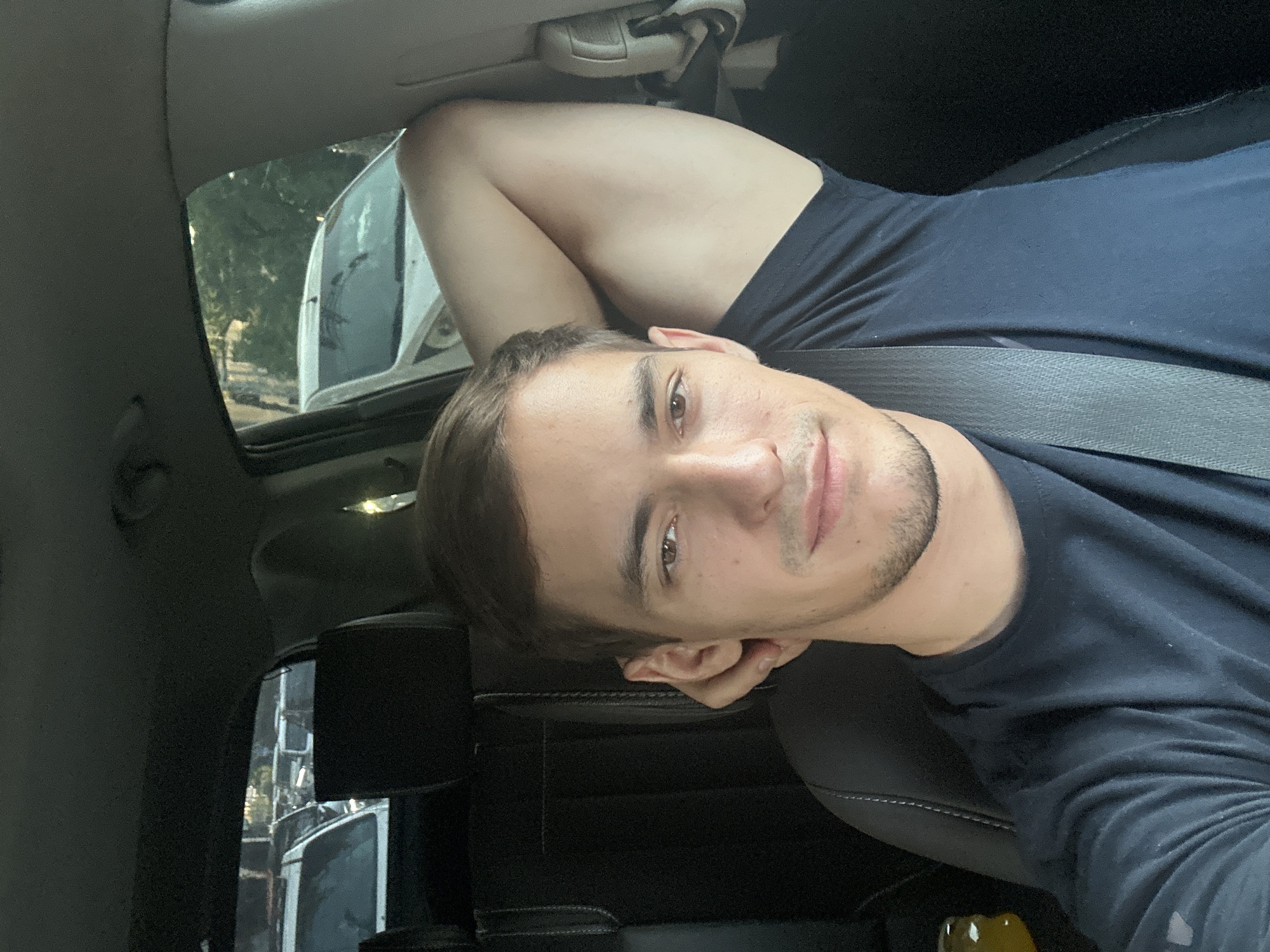 A man is seated in the driver's seat of a car, looking towards the camera. His right arm is bent and resting behind his head, with his hand touching his left ear. He is wearing a dark blue t-shirt and a grey seatbelt is visible across his chest. The interior of the car, including black leather seats, is visible. Outside the car, other vehicles and trees can be seen through the windows. The lighting suggests it is daytime. The image is a selfie taken from inside the car. The specific location in the city of Tashkent, Uzbekistan is not identifiable from the visual information present in the image.