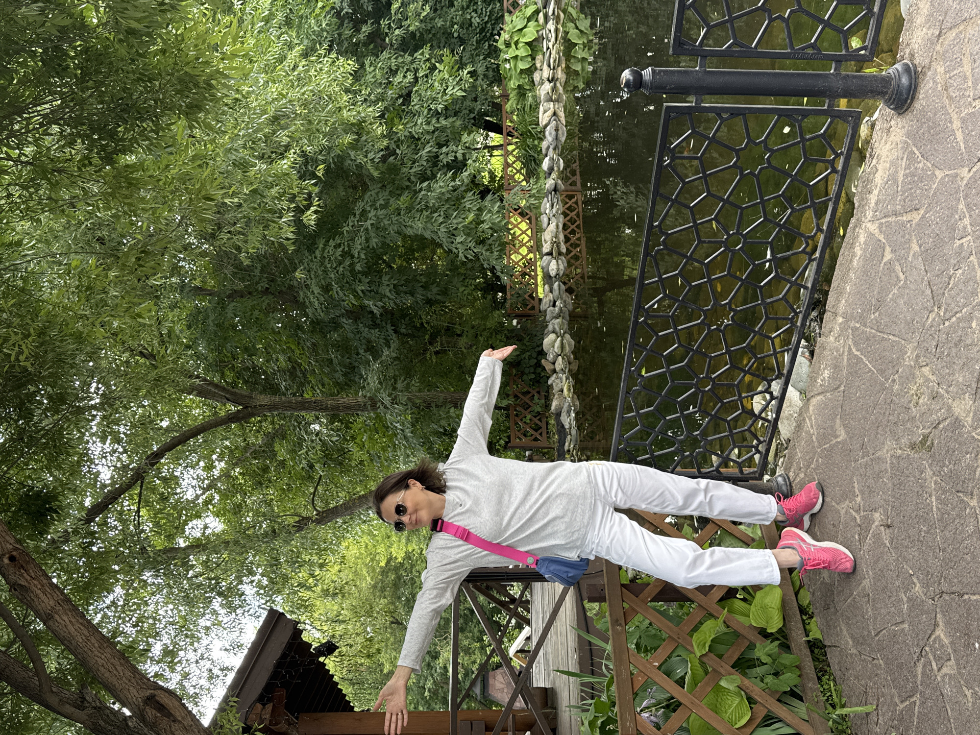 A woman stands outdoors on a paved walkway with her arms outstretched. She is wearing white pants, a light grey long-sleeved shirt, pink sneakers, sunglasses, and a pink and blue crossbody bag. Behind her is a pond with a decorative black metal fence along its edge. A wooden fence and a building with a dark roof are to her left. Lush green trees and foliage fill the background. The scene appears to be a park or garden. The location is the Izhevsk Zoo in Izhevsk, Russia.