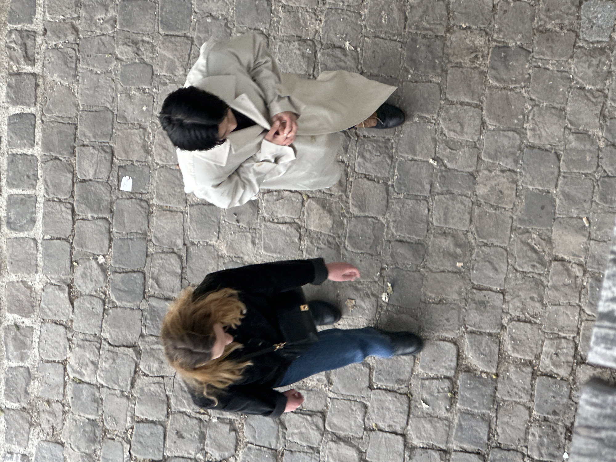 The image shows an overhead perspective of two individuals on a cobblestone street. One person is wearing a light-colored trench coat and appears to be standing still with their hands clasped in front of them. The other person is wearing a black coat, blue jeans, and a purse or bag across their body, appearing to be walking. The cobblestone pavement is worn. Based on the cobblestone street and architecture style, the location is likely in Paris, France.