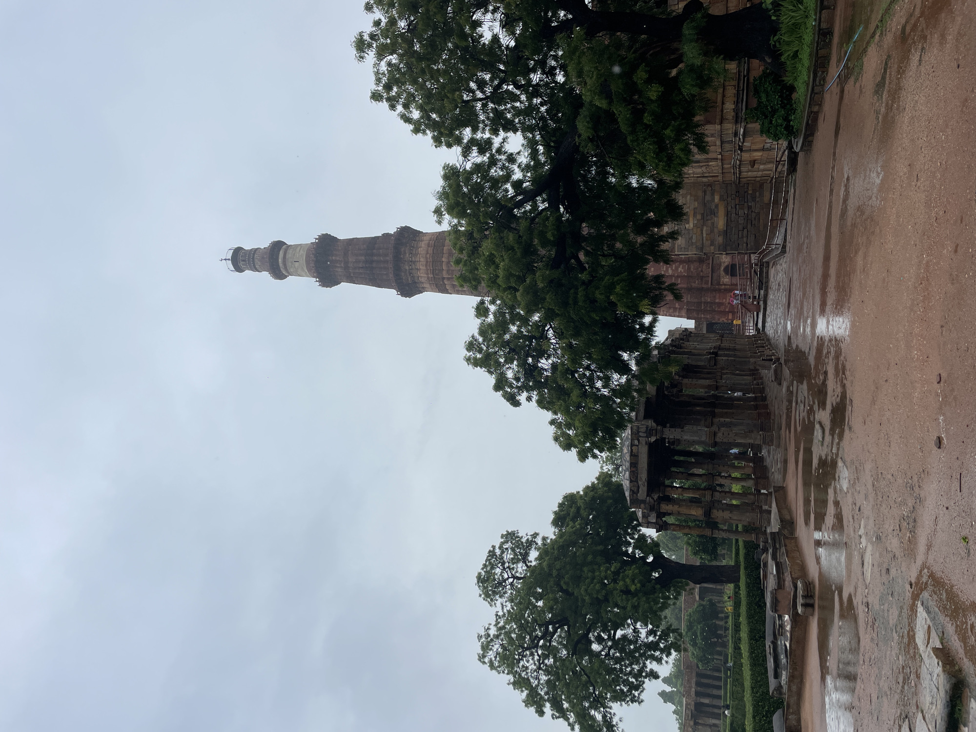 The image depicts the Qutub Minar complex in Delhi, India, under an overcast sky, likely after rainfall, as evidenced by wet ground and puddles. The dominant subject is the Qutub Minar, a towering minaret constructed of red sandstone, with multiple tiers and intricate architectural details, prominently visible in the background. In the foreground, a partially ruined stone structure, possibly a pavilion or colonnade with numerous stone pillars, stands to the left. Lush green trees, some with dense foliage, are scattered throughout the scene, partially obscuring views of the minaret and surrounding historical structures. The ground is a mix of red earth and stone paving, wet and reflective in places. A few individuals are present in the distance, near the base of the minaret and within the complex, some appearing to hold umbrellas, suggesting ongoing or recent precipitation. The overall atmosphere is somber and damp, with muted colors due to the cloudy weather.