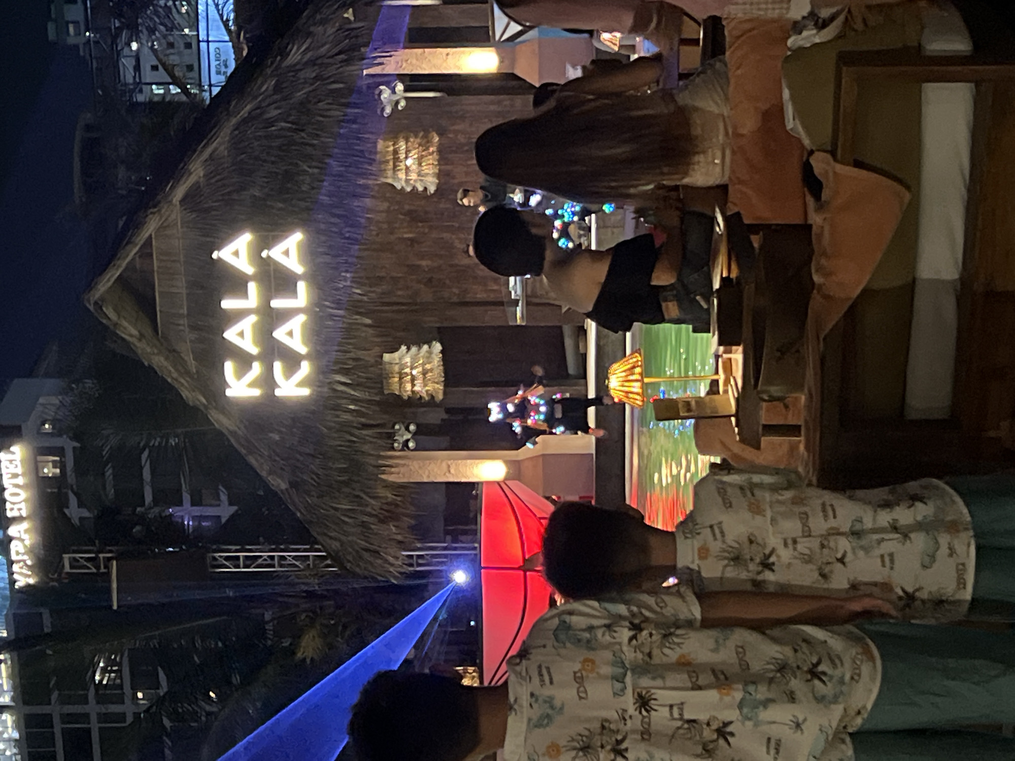 The image depicts an outdoor evening scene at a venue in Đà Nẵng, Vietnam. The prominent feature is a thatched-roof structure with illuminated neon signs spelling "KALA KALA". In the foreground, two young boys are viewed from behind, both wearing patterned shirts and teal pants. To the right, a group of people are seated at tables, suggesting a dining or lounge area. Behind them, a performance is taking place by a pool, featuring a person in a costume illuminated with colorful lights and laser projections. In the background, the "YAPA HOTEL" sign is visible on a multi-story building. The overall ambiance suggests a lively entertainment or dining establishment.