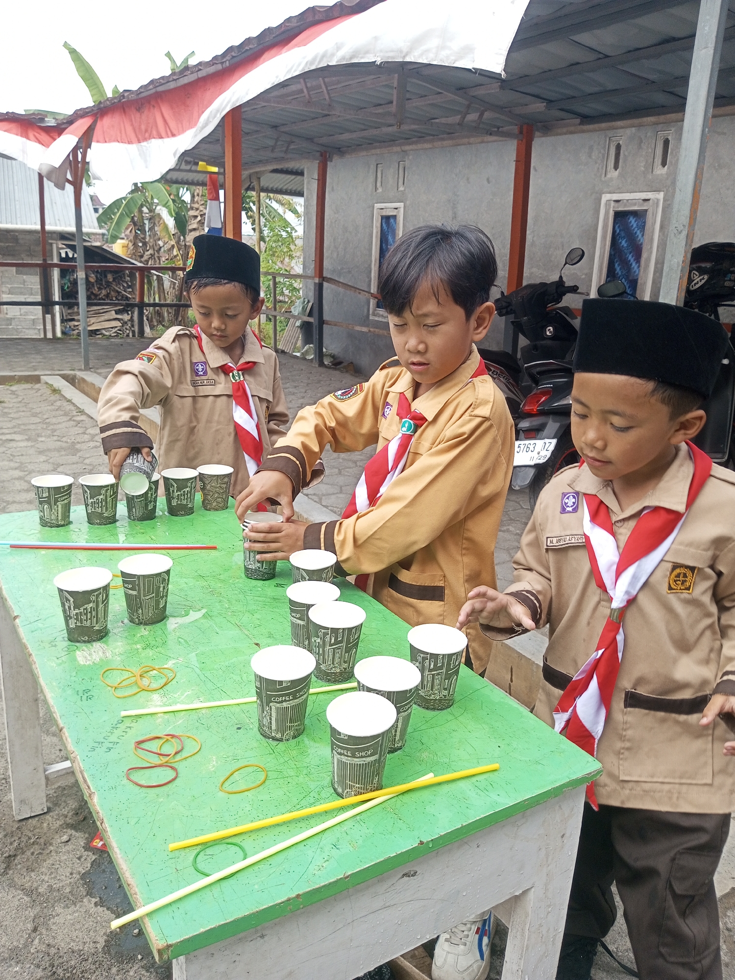 Three young boys, dressed in brown scout uniforms with red and white neckerchiefs and black caps, are gathered around a green table outdoors. The table is covered with paper cups, rubber bands, and yellow straws. The boy on the left is pouring liquid from a cup into another cup on the table. The boy in the center is also pouring liquid from a cup into a cup on the table. The boy on the right is placing a cup onto the table. In the background, there is a motorcycle, a building with grey walls and windows, and trees. A red and white Indonesian flag is visible on the roof structure. The scene appears to be in Kalibeber, Indonesia, likely during an outdoor activity or event.