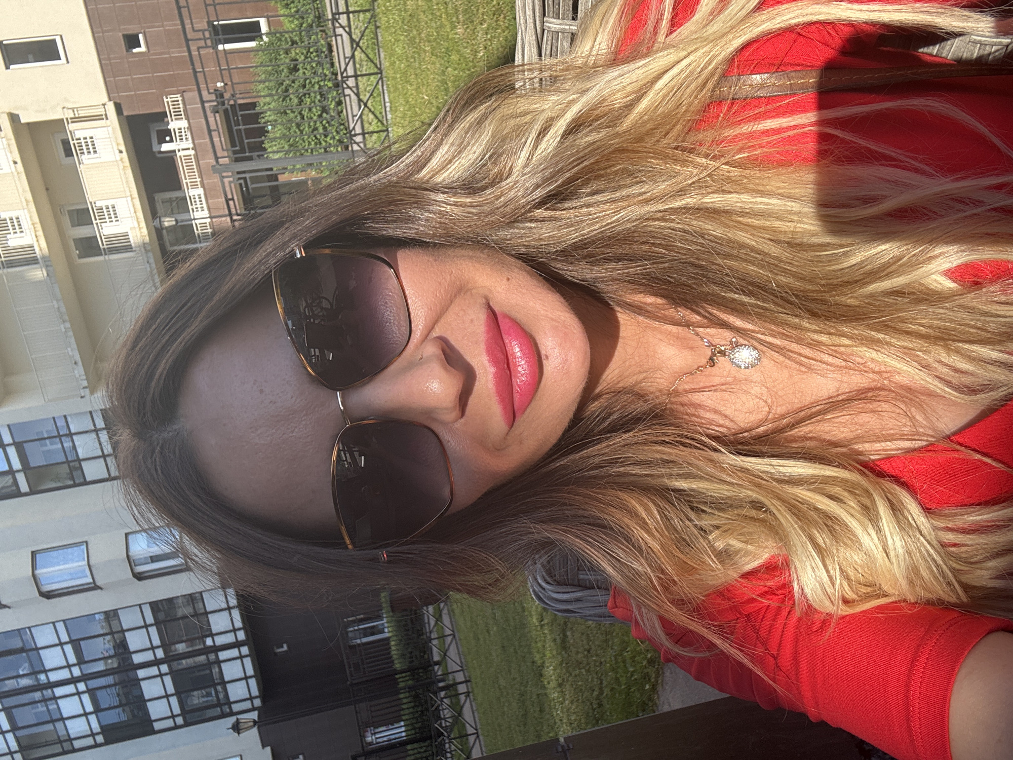 The image is a close-up, selfie-style shot of a woman with blonde hair, wearing sunglasses and a red top. Her lips are painted with a vibrant pink lipstick. She is wearing a delicate necklace with a pendant. The background is blurred, but appears to be an outdoor setting with buildings and green grass, suggesting it is daytime. The lighting indicates it's a sunny day. The woman's expression is neutral. The scene suggests a casual outdoor moment, possibly in a residential area given the buildings visible in the background.