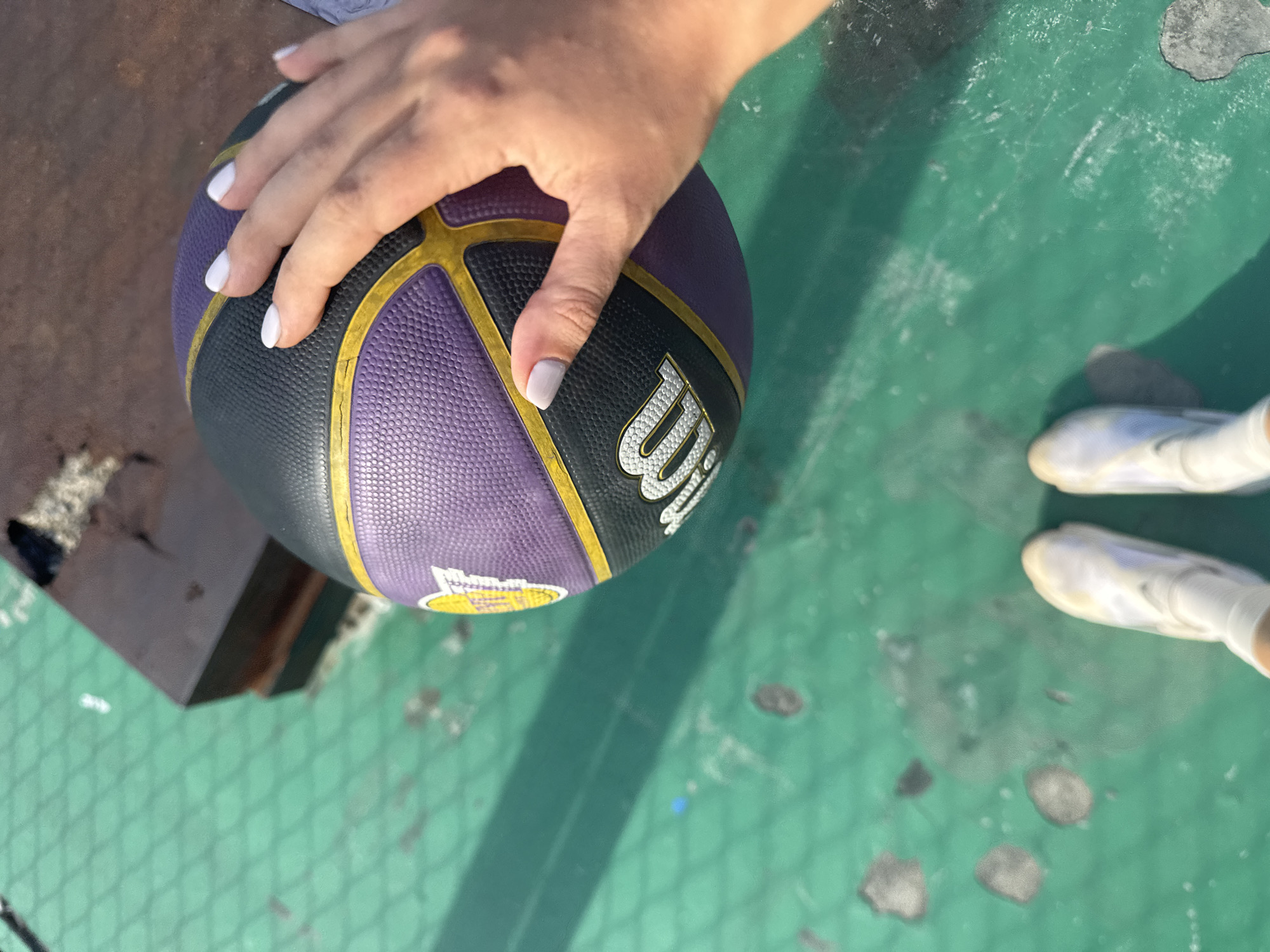 A first-person perspective shot shows a person's left hand resting on a purple and black basketball with yellow trim. The basketball is on a green, textured surface. The person's feet, clad in white sneakers, are visible at the bottom of the frame, also positioned on the green surface. In the background, to the left of the basketball, is a dark, rectangular object. The green surface appears to be outdoors, possibly a basketball court, with some debris and discoloration visible. The lighting suggests it is daytime, with shadows cast across the surface. The location is not explicitly stated as being in Ko Samui District, Thailand.