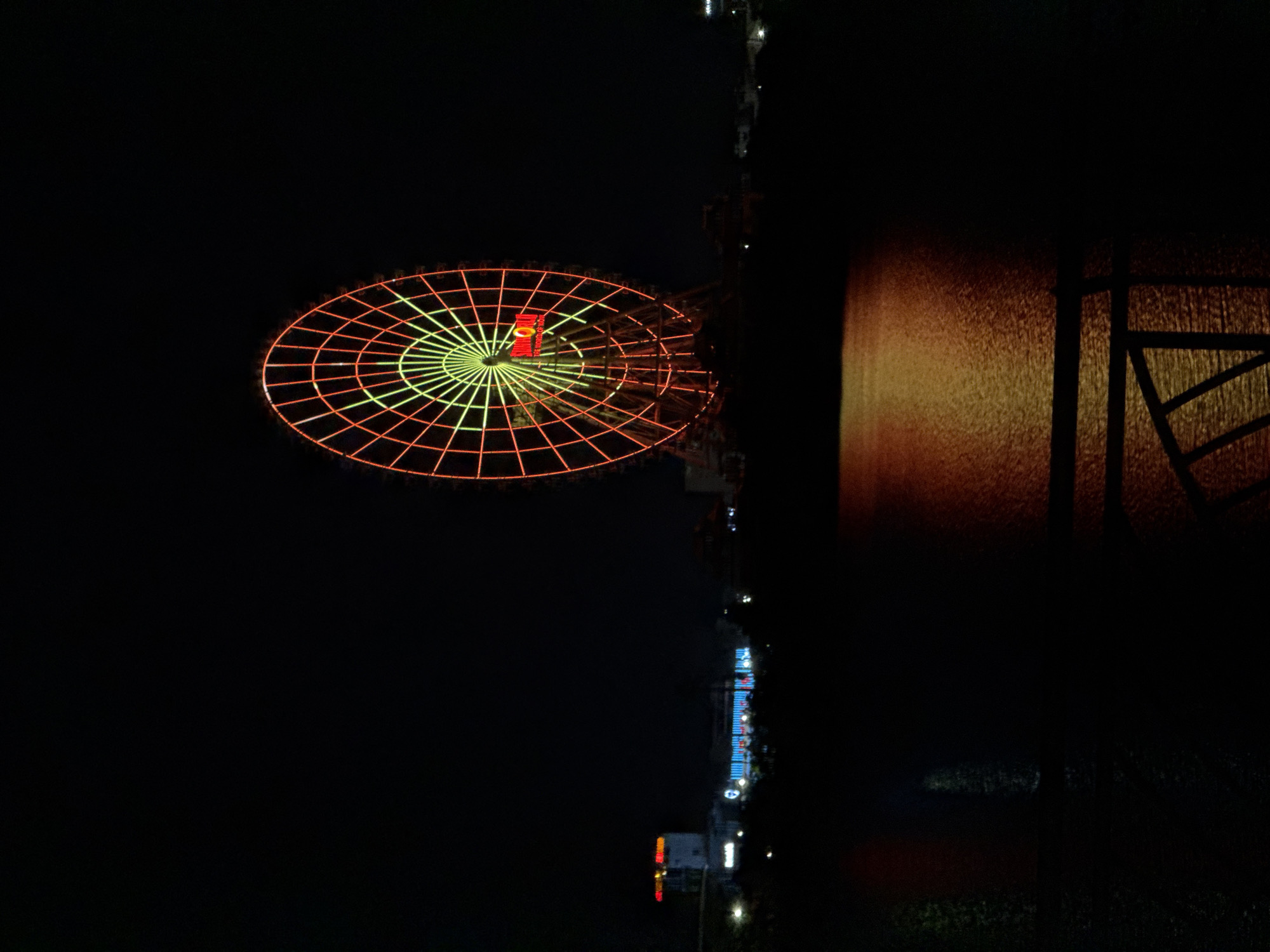 At night, a Ferris wheel is visible in Đà Nẵng, Vietnam. The wheel is illuminated with yellow and red lights. The ground is very dark but shows signs of a road, a water source, and more lit-up buildings. The image is oriented horizontally in a way that makes the horizon line go vertically.