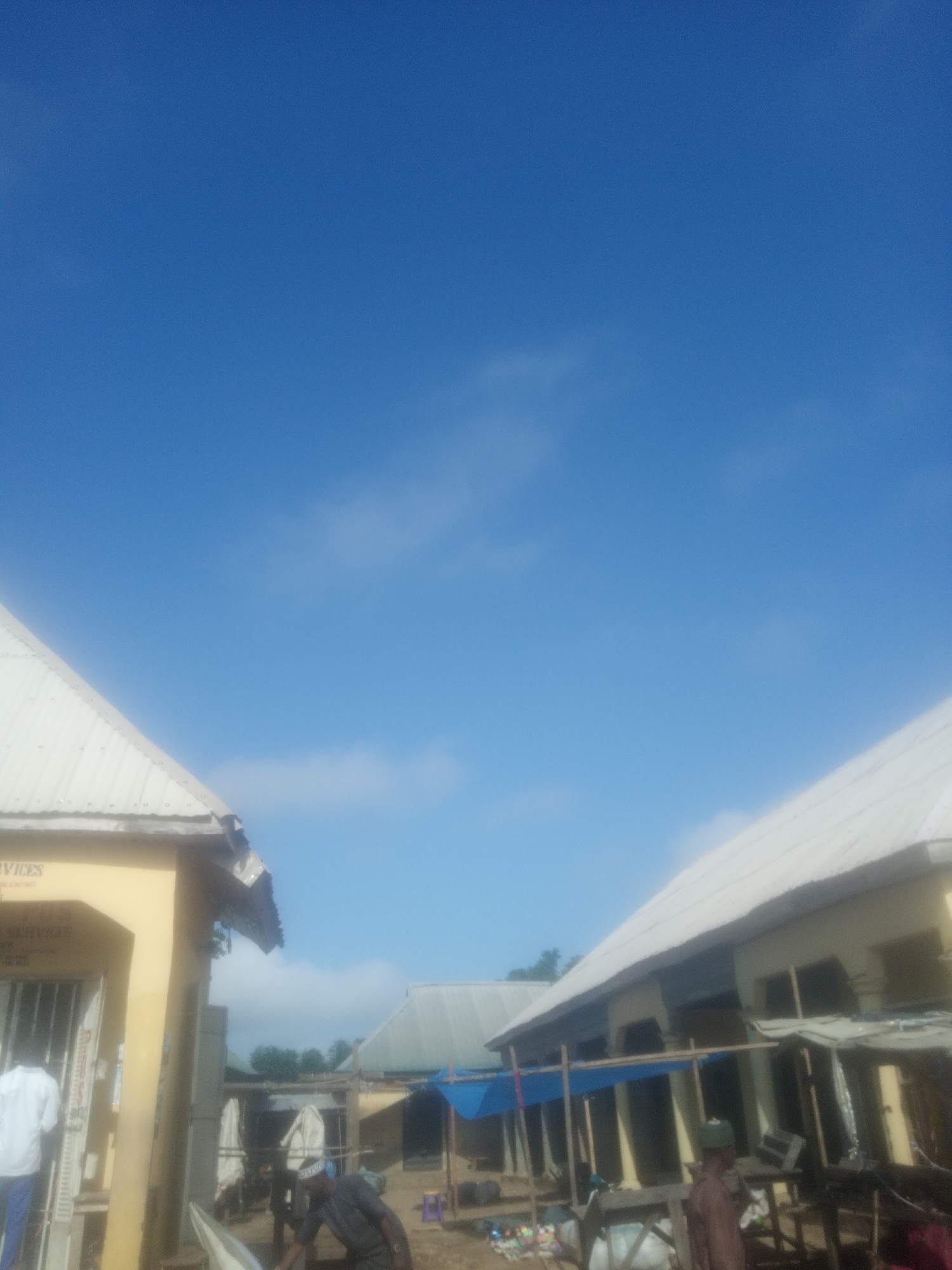 In Serti, Nigeria, an outdoor scene unfolds under a vast, clear blue sky with scattered wispy clouds. The foreground is dominated by the corner of a yellow building with a corrugated metal roof. Visible text on the building reads "SERVICES" and "GENERAL." To the left, a person in a white shirt and blue pants stands facing away from the camera.
Further into the image, another building with a similar corrugated metal roof stretches diagonally. In front of this structure, a series of stalls or awnings are set up. A blue tarp covers part of this area. Several people are present. One individual, wearing a dark cap and tunic, is partially visible in the lower right. Other individuals are also present but their details are less clear. Various objects, including what appear to be goods or supplies, are scattered around the market area, and a blue plastic stool is visible. The scene suggests a bustling market or commercial area in the town.