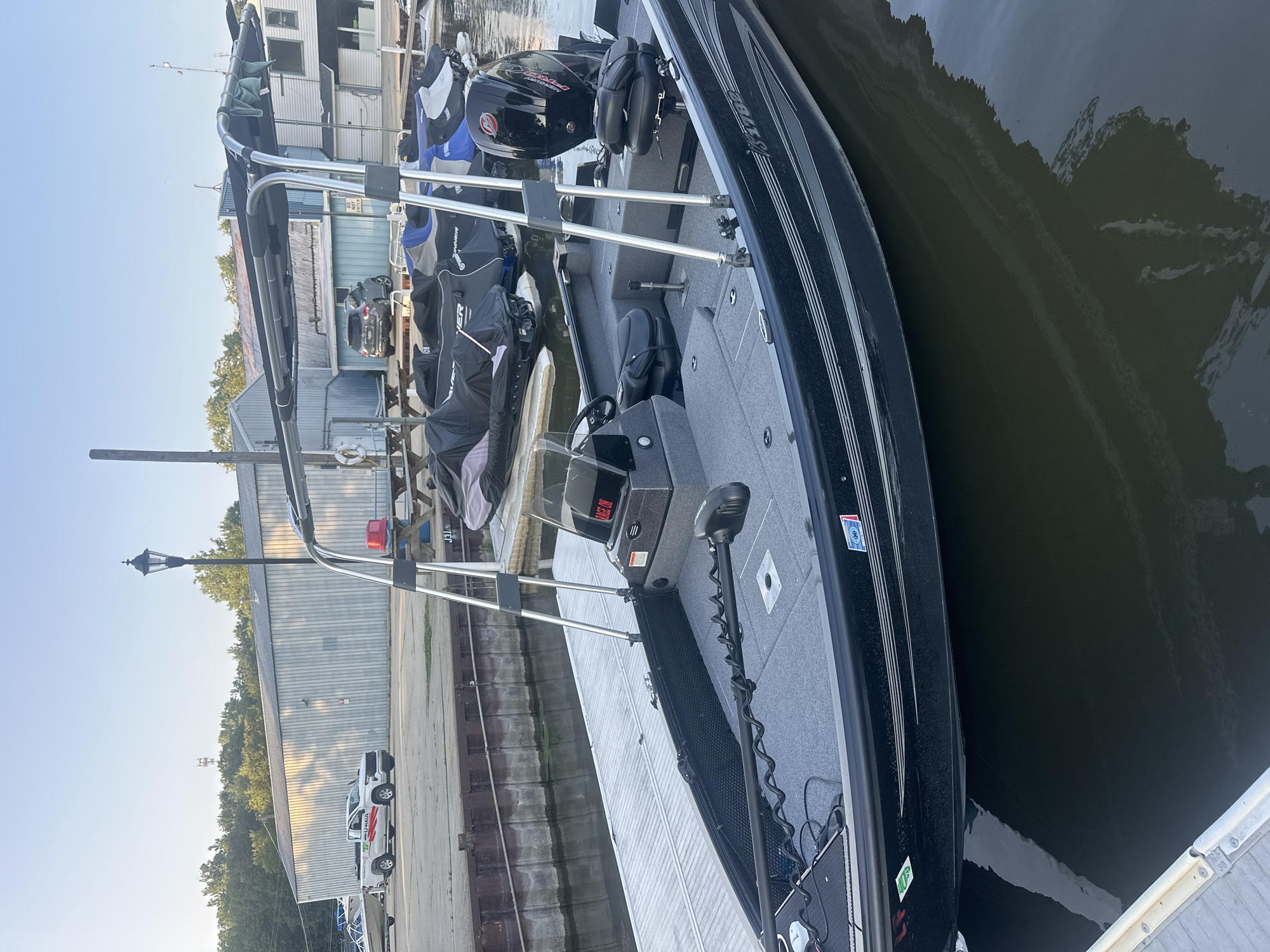 An exterior, eye-level, medium shot shows a dark blue fishing boat docked at a marina on a bright, sunny day. The boat is positioned facing to the right, with its bow to the left of the frame. The boat has a dark gray interior with two bucket seats, a steering console, and a trolling motor mounted on the bow. A Bimini top with a dark gray fabric is extended over the cockpit area, supported by a silver metal frame. Several jet skis covered with dark gray tarps are docked to the right of the fishing boat. In the background, a U-Haul pickup truck is parked near a large, light-colored industrial building with a gray metal exterior. A street lamp with a black lantern is visible behind the truck. Trees line the horizon behind the building. The water in the marina is calm with slight reflections. The scene is located in the city of Algonquin, United States of America.