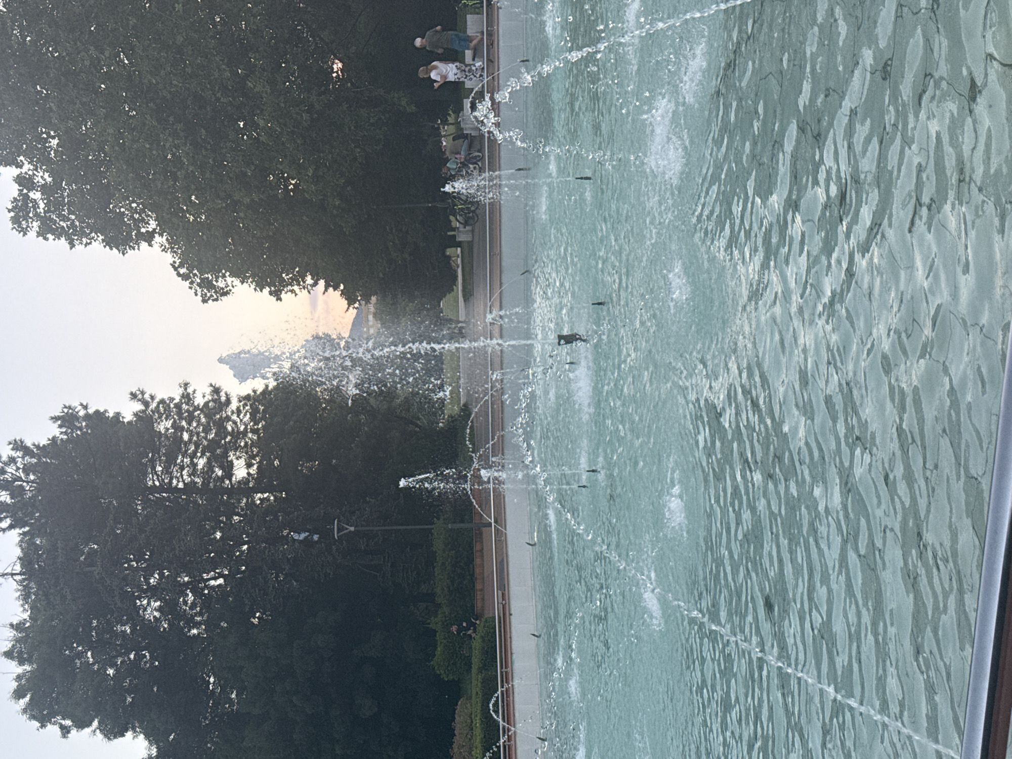 This media file depicts a fountain with water jets spraying upwards in what appears to be a park setting. The water is a turquoise color and reflects the surrounding environment. In the background, there are lush green trees, some of which are quite tall and dense. A pathway runs alongside the fountain, and on this path, a few people are visible. One group of two individuals, possibly adults, are standing near the edge of the fountain. Further down the path, several bicycles are parked, suggesting that people have arrived by bicycle. The overall scene is tranquil and suggests a pleasant outdoor environment. The lighting indicates it is likely daytime, possibly late afternoon or early evening, as the light is somewhat diffused and soft. The sky in the distance appears bright. The presence of trees and a well-maintained pathway points to a public park or garden. The location is identified as Sofia, Bulgaria. There is no visible text in the image.