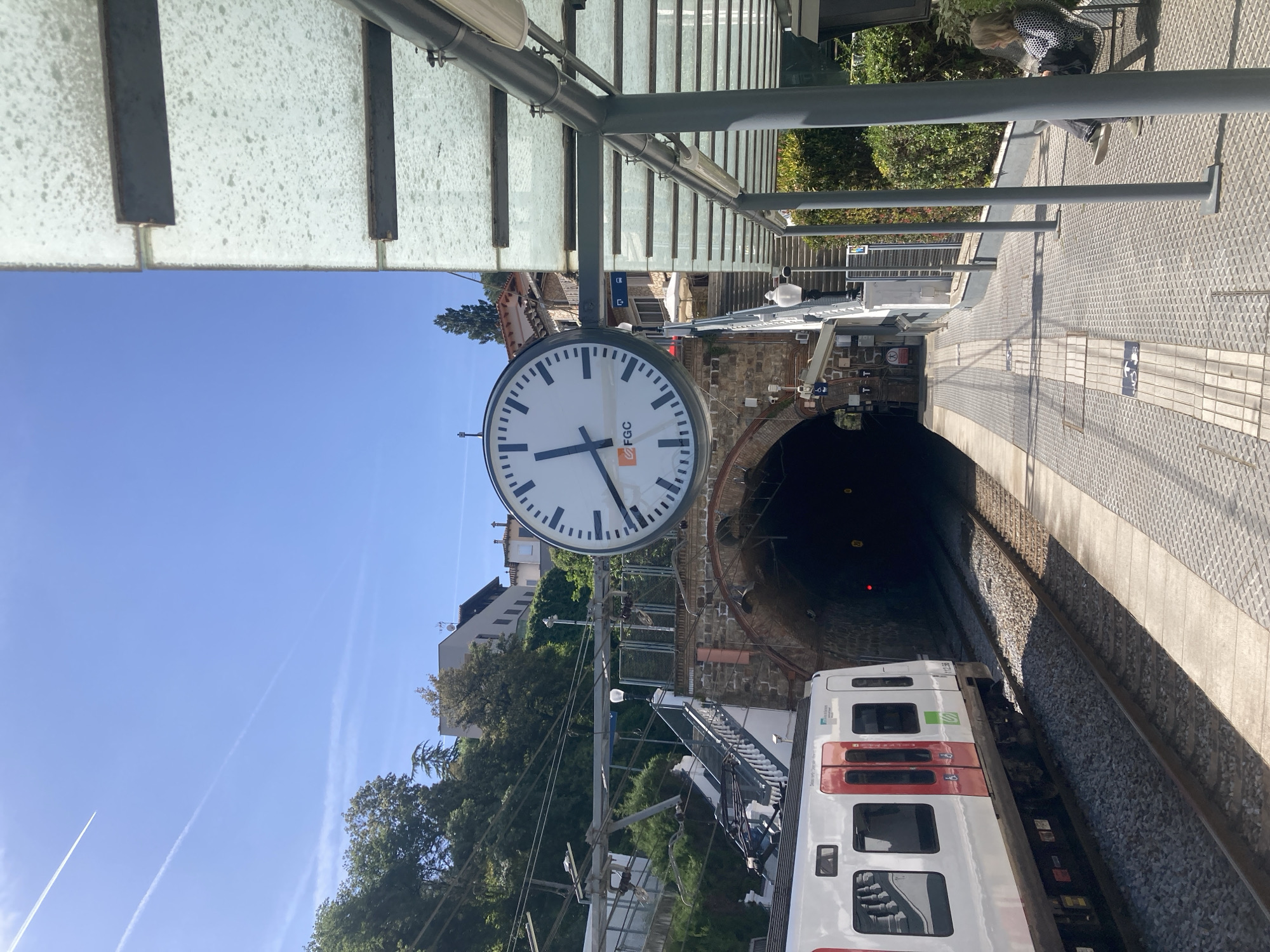 This photograph depicts a train station platform with a clock mounted overhead. The clock has a white face with black numerals and hands, and a logo that reads "FGC". In the background, a train is entering a tunnel. The platform is constructed of a textured gray material, and a shelter with a metal frame and glass roof provides cover. Vegetation is visible behind the tunnel entrance, and buildings can be seen in the distance. The sky is clear and blue. A person is sitting on a bench on the right side of the image.