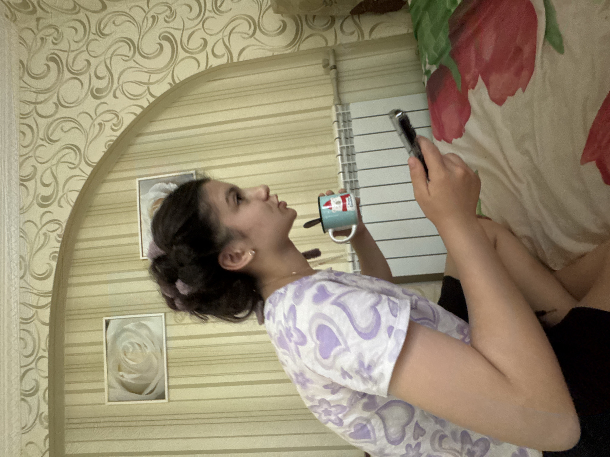 A young woman with dark hair pulled back in a bun, wearing a white t-shirt with purple hearts, sits on a bed. She is holding a mug in her right hand and a phone in her left hand. The mug has a picture of Santa Claus on it. The bed is covered with a comforter that has a floral pattern of red and green flowers. Behind her, a white radiator is visible. The wall is covered with striped wallpaper and decorated with a framed picture of a white rose. The scene appears to be indoors, likely in a bedroom.