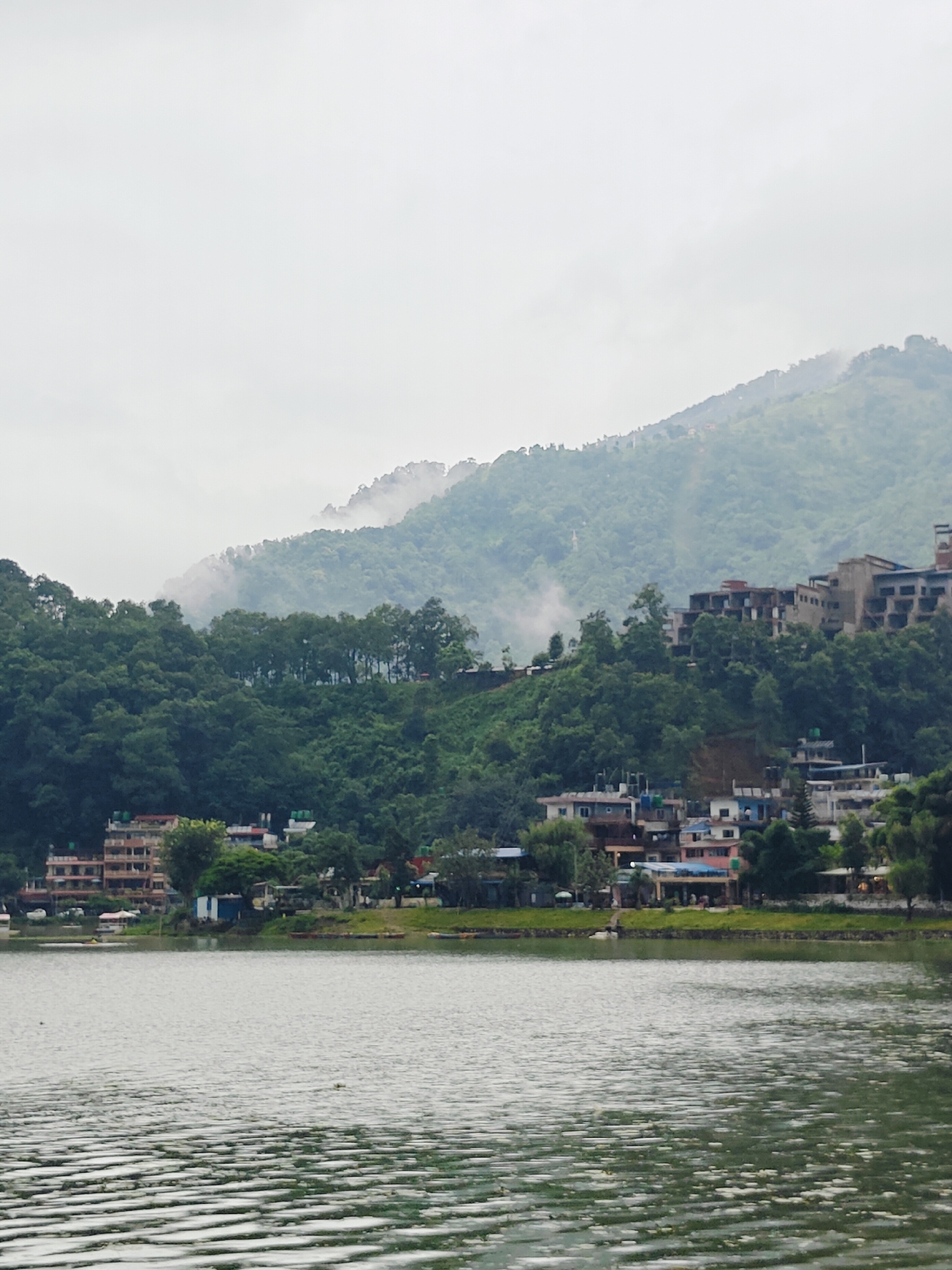 This image depicts a scenic view of Phewa Lake in Pokhara, Nepal. The foreground is dominated by the lake's surface, which is rippled and reflects the overcast sky. In the midground, the shore of the lake is lined with buildings and lush green vegetation. Several boats are visible docked or on the water. Behind the shoreline, a hillside is densely covered with trees, and further back, a larger mountain looms, partially shrouded in mist and fog. Buildings are visible nestled within the greenery on the hillside. The overall atmosphere is serene and tranquil, characteristic of a cloudy day in the Himalayan region.