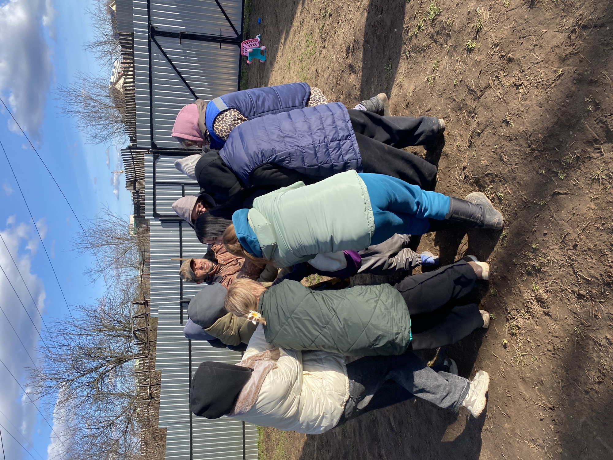 The image shows a group of approximately eight people lying on the ground in what appears to be a muddy, dirt area. The location is Chaikovsky, Russia. The people are lying in a line, one on top of the other. They are all wearing winter clothing, including jackets, hats, and boots. The background includes a corrugated metal fence, trees, and a blue sky with clouds. In the background is also a pink shopping cart. The individuals are arranged in a way that suggests they are sheltering from potential danger.