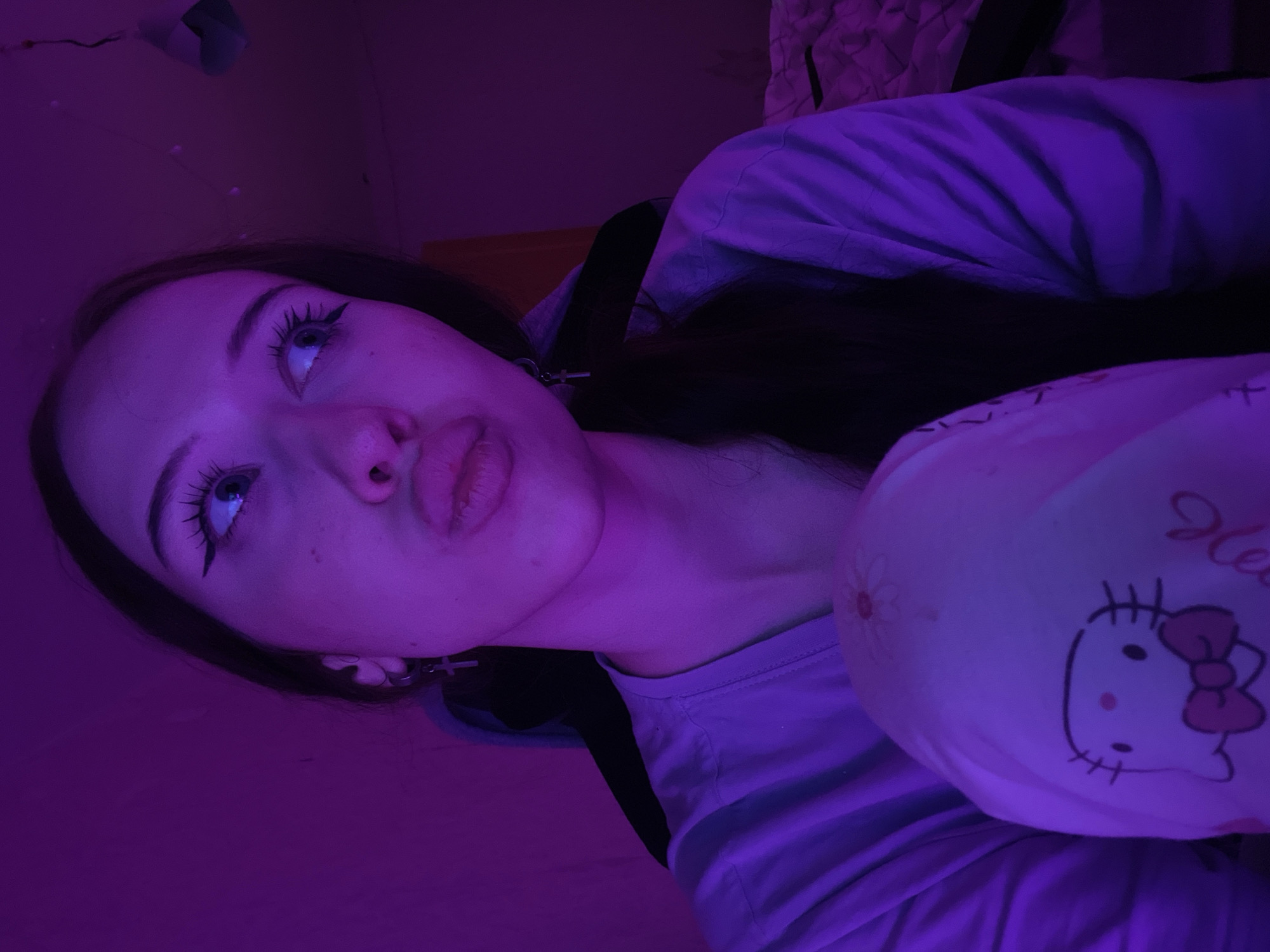 This is a vertically oriented, close-up, indoor photograph of a young woman bathed in purple light. The woman is lying down, with her head tilted upwards and to the left. Her eyes are wide open, looking upwards, and her eyelashes are heavily emphasized with mascara. She has a slight pout to her lips. She is wearing a light-colored t-shirt and appears to be wearing a choker necklace with a small cross pendant.
To the right of the woman is a pillow with a Hello Kitty design. The Hello Kitty character is visible on the pillow, complete with her signature bow. The background is dark and out of focus, with some texture suggesting a wall or bedding. The purple lighting creates a moody and atmospheric effect.
The time of day is not discernible from the image, nor is the weather. The presence of bedding and the intimate framing suggest a bedroom. The overall emotion conveyed is one of introspection or a playful pose. No visible text is present in the image other than the "Hello" written on the Hello Kitty pillow. The location context of Votkinsk, Russia, is provided externally and does not offer any specific visual cues within the photograph.