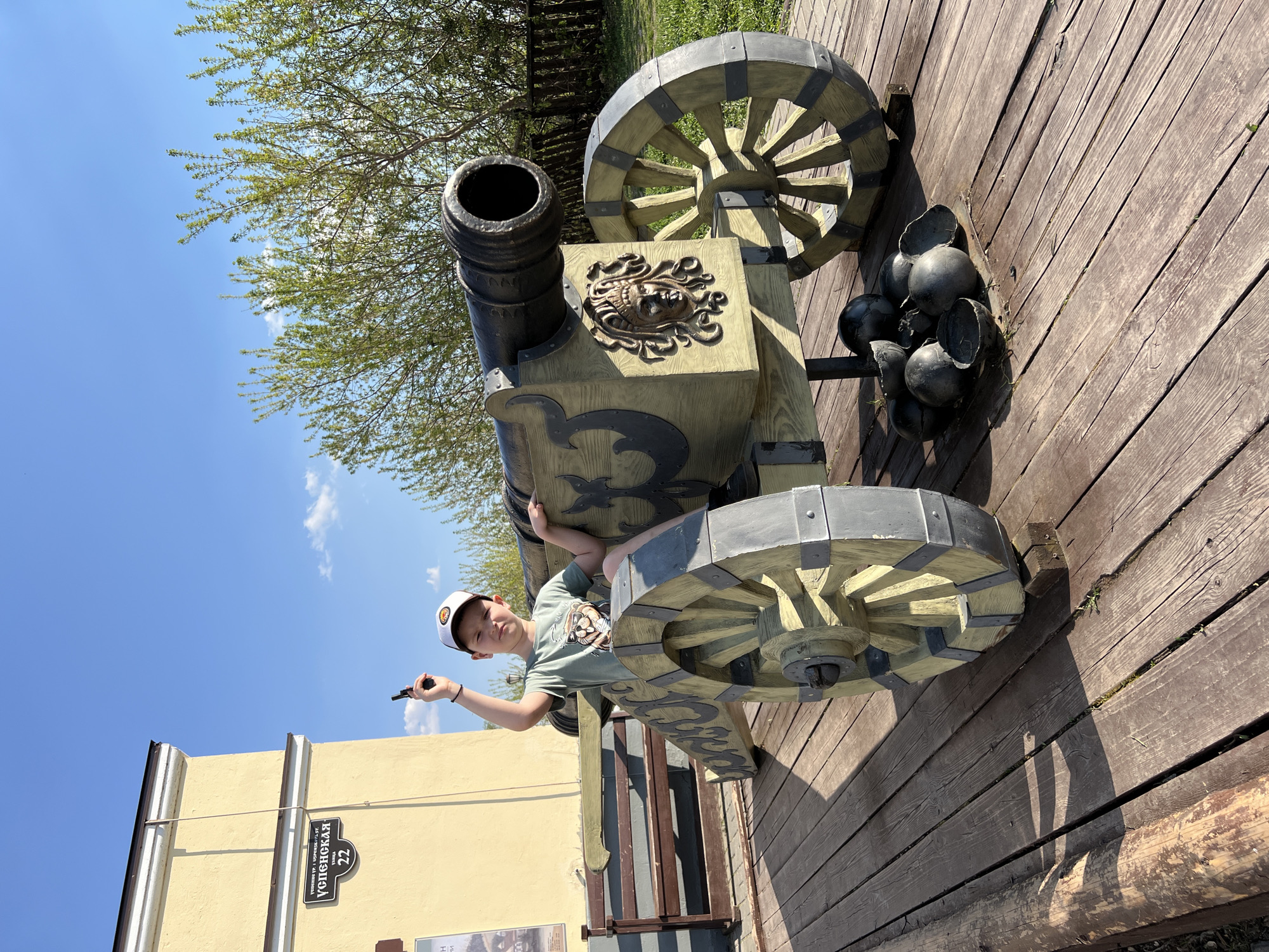 A young boy wearing a light green shirt and a baseball cap is straddling a large, decorative cannon on a wooden deck. He is holding a small black object in his right hand, pointed upwards. The cannon is painted in shades of gray and yellow, with decorative metal accents. To the right of the cannon are cannonballs. In the background, there is a yellow building with a sign that reads "Успенская 22", indicating the location is Uspenskaya Street, number 22. A tree with green leaves is partially visible in the upper background, against a blue sky with some clouds. The scene appears to be in Sviyazhsk, Russia, based on the street name written in Russian.