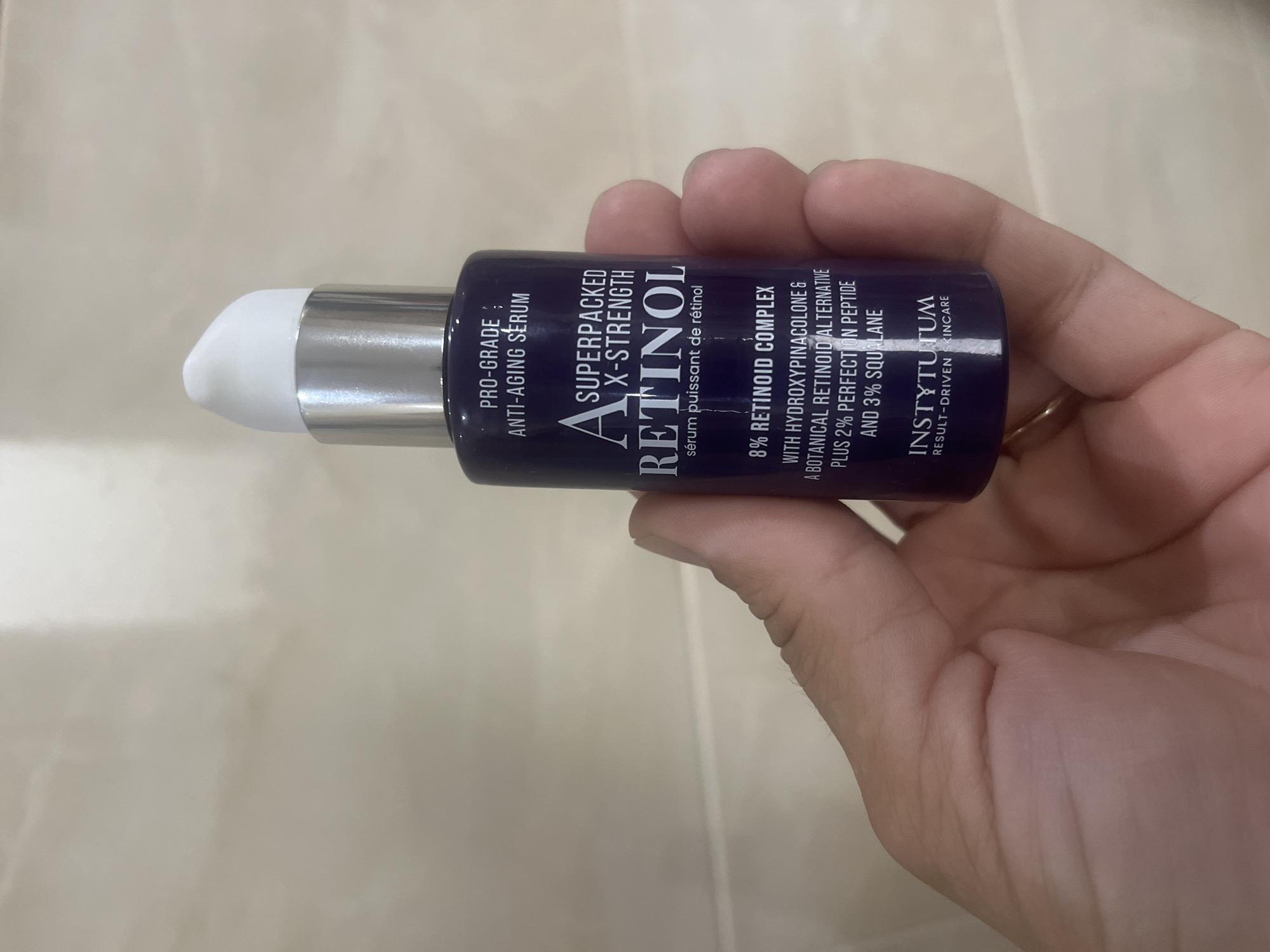 A person's hand holds a dark purple bottle of anti-aging serum. The bottle has a silver pump dispenser with a white cap. The label on the bottle reads "PRO-GRADE ANTI-AGING SERUM," "A SUPERPACKED X-STRENGTH RETINOL," and "INSTYTUTUM RESULT-DRIVEN SKINCARE." The background is a textured wall that appears to be light beige or cream colored. The image does not provide any information about the location in the city of Onokivtsi, Ukraine.