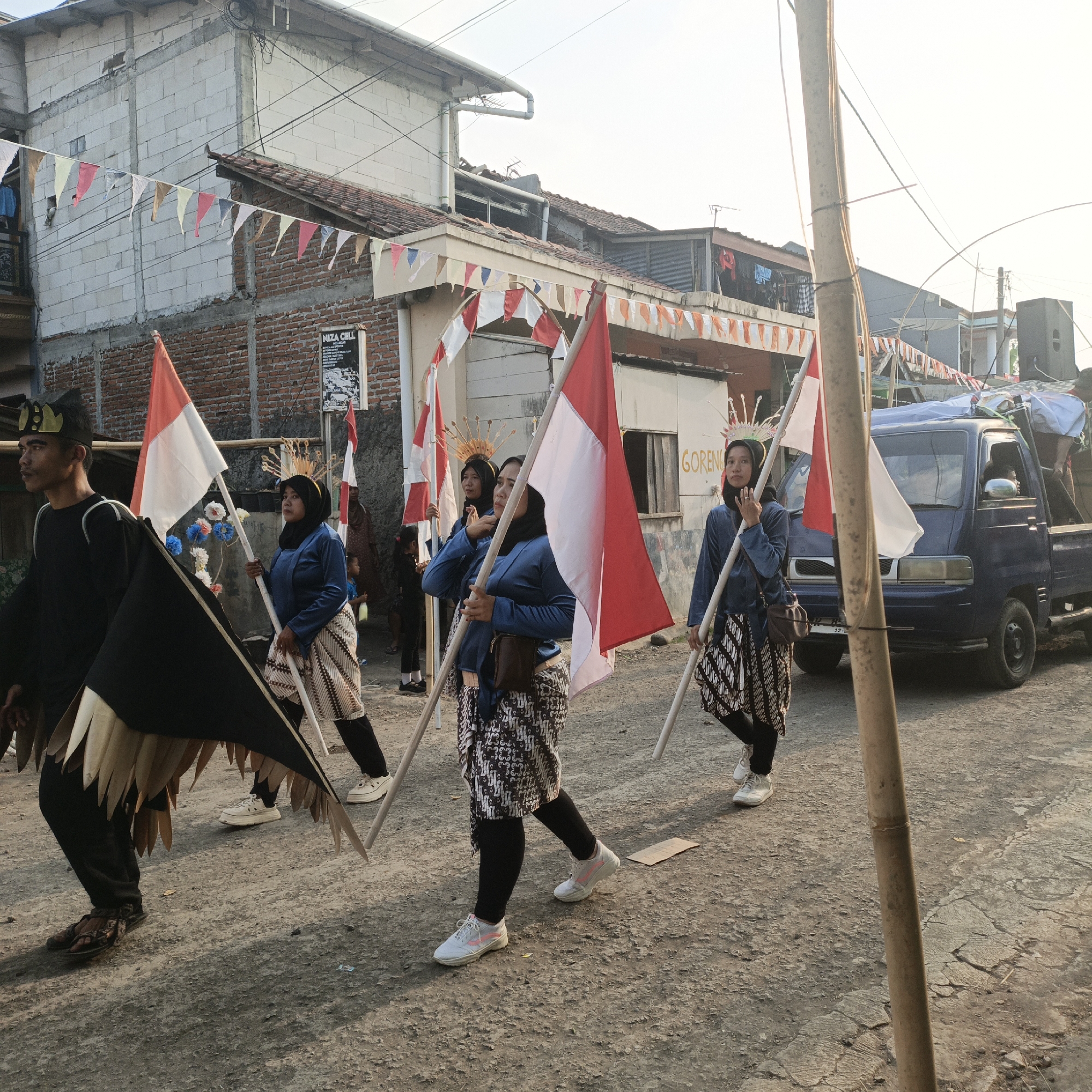 In Tanahbaya, Indonesia, a group of individuals, appearing to be participants in a parade or cultural event, walk down a paved street. The foreground is dominated by the lower bodies and legs of these individuals. One person on the left wears black pants and brown sandals, with a black and gold headdress partially visible. Another individual in the center, also wearing black pants and white sneakers with pink accents, holds a red and white flag. A person to the right, dressed in a blue long-sleeved shirt and a patterned sarong over black leggings, holds a similar red and white flag. The flags are Indonesian national flags. Behind them, a blue pickup truck is parked on the right side of the road, and a blue speaker is visible on its bed. The background shows buildings constructed from brick and concrete blocks, with red tile roofs. Numerous colorful triangular flags are strung across the street overhead, adding to the festive atmosphere. The lighting suggests it is either early morning or late afternoon, with the sun casting long shadows.