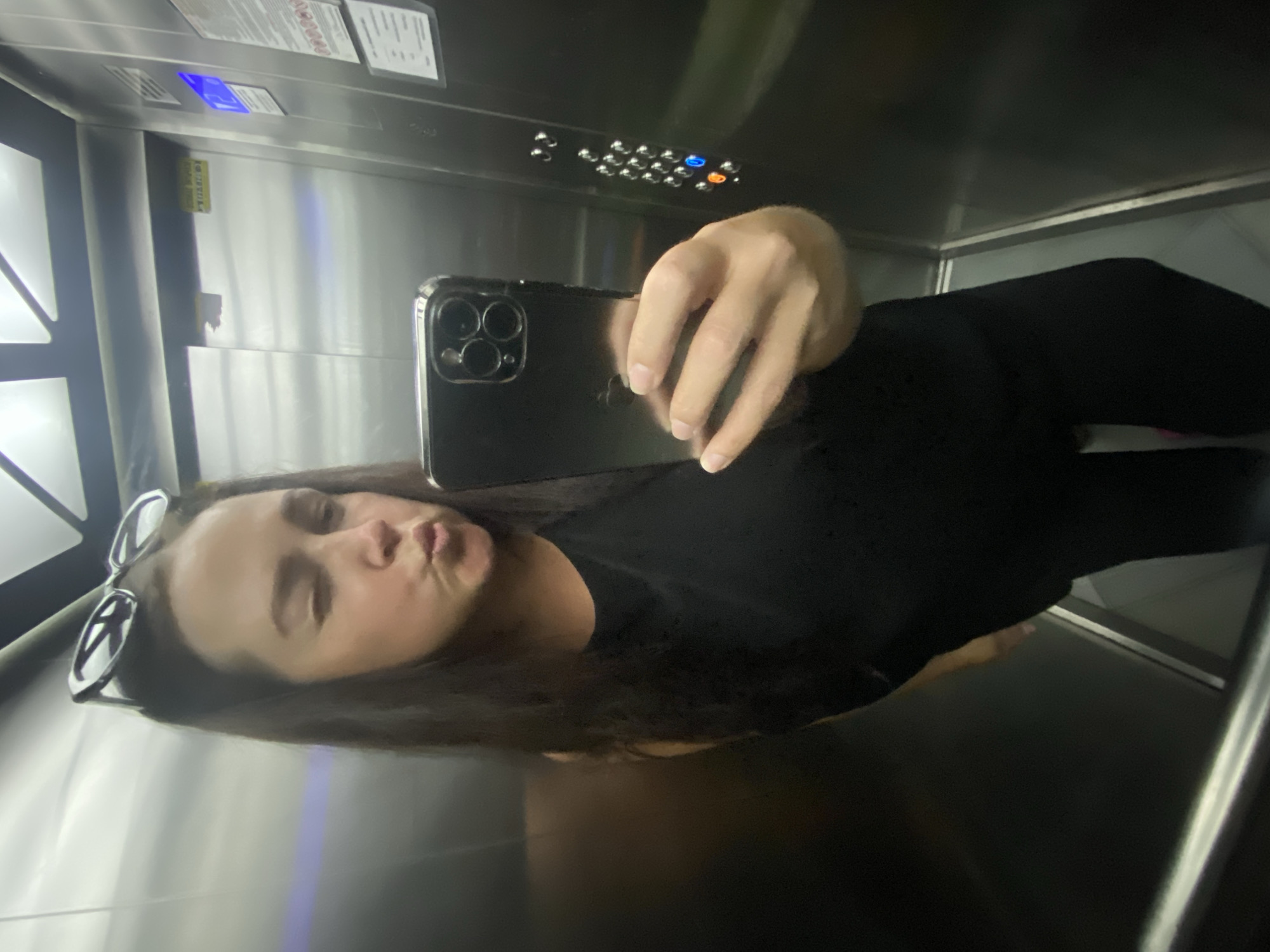 A woman is taking a selfie in an elevator. She is wearing a black tank top and black leggings, and she has sunglasses on top of her head. She is holding an iPhone in her right hand, and she is making a kissing face at the camera. The elevator is made of stainless steel and has a panel of buttons on the right side. There is a blue light on the panel, indicating that the elevator is in motion. The location is the city of Sochi, Russia.