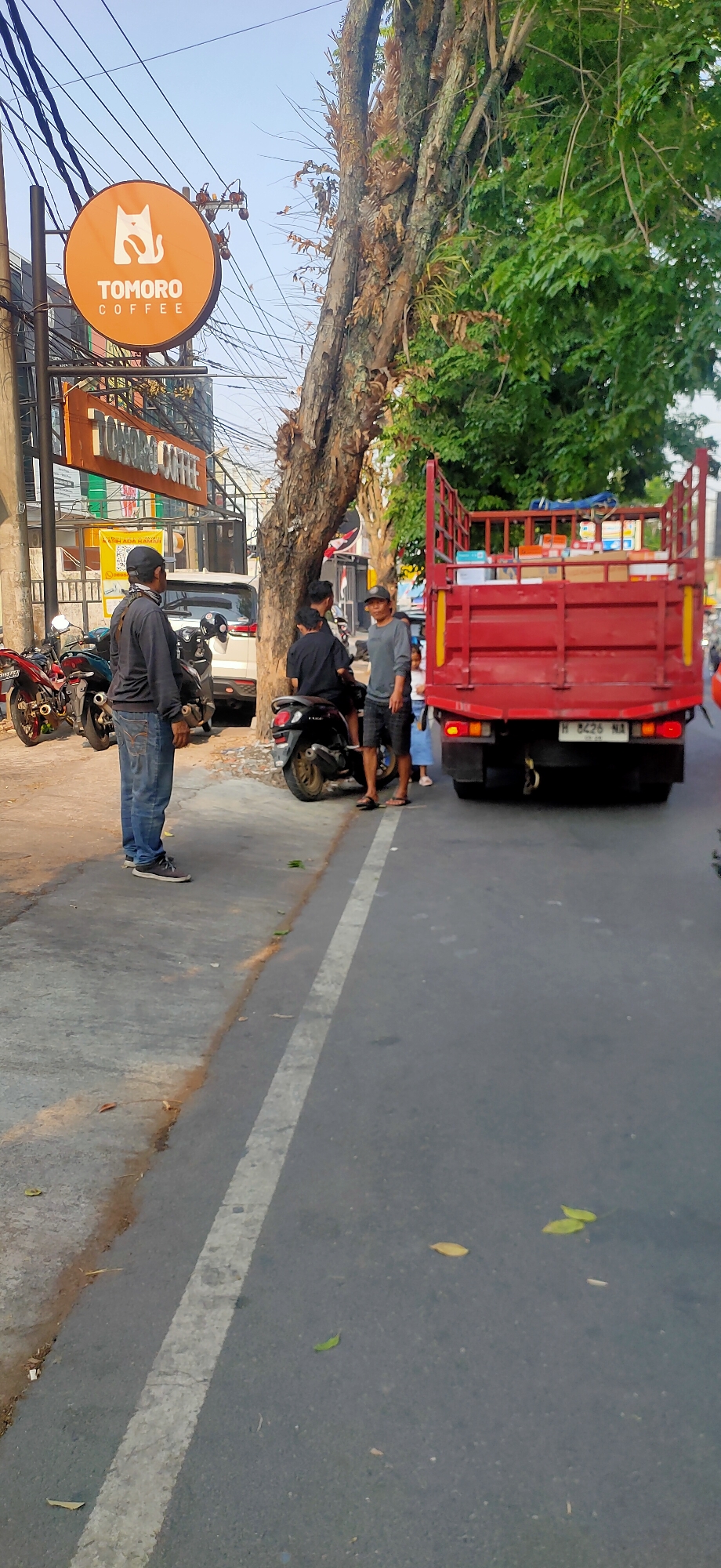 This is a medium shot, taken from a slightly low angle, of a street scene in Semarang, Indonesia, featuring a red truck, several people, motorcycles, and a cafe sign. The sun is shining, casting shadows and illuminating the scene.
In the foreground, to the left, is a man wearing a dark long-sleeved shirt, blue jeans, and a black baseball cap. He is standing on a sidewalk next to a parked motorcycle. Behind him, another motorcycle is parked.
In the middle ground, a red truck with its bed loaded with boxes is parked on the street. Three men are standing near the truck, with two of them interacting with each other. One man, wearing a gray long-sleeved shirt and shorts, is looking towards the camera. Another man, in a black shirt and black pants, is sitting on a motorcycle. A third man, also in a black shirt, is standing next to the motorcycle. A young girl in a light blue dress is partially visible near the truck.
To the left of the scene, a bright orange circular sign with a white cat silhouette and the text "TOMORO COFFEE" is visible, mounted on a pole. Below it, a horizontal sign with the same "TOMORO COFFEE" text is also present. Several power lines crisscross the sky above. A large tree with rough bark is prominently featured to the right of the cafe sign, with lush green foliage.
The street is paved and has a white line marking the lane. Leaves are scattered on the sidewalk and the road. The background shows buildings and more trees. The overall impression is a bustling street scene with commercial activity.