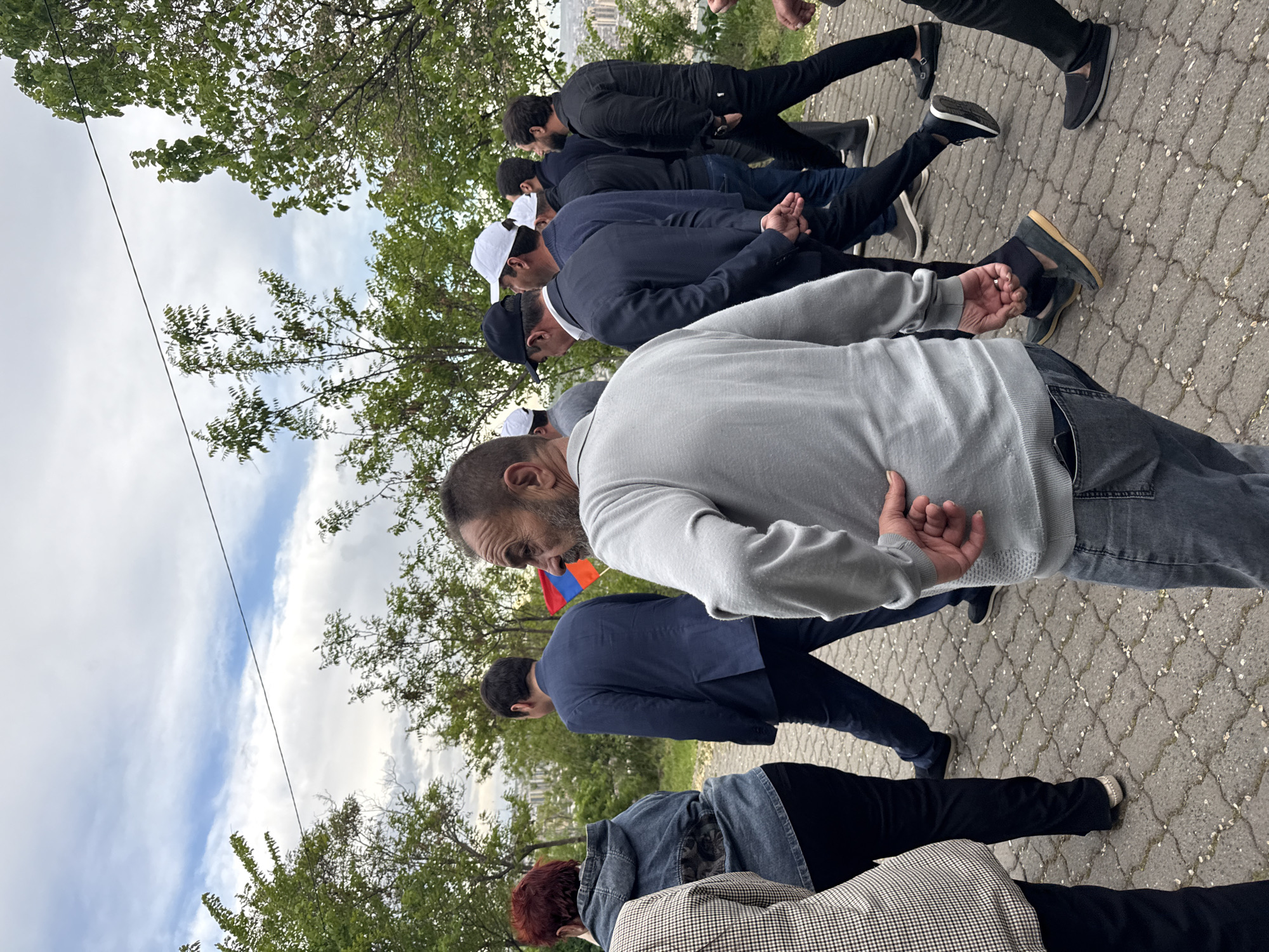 A group of people is walking outside on a paved pathway lined by trees, in what appears to be the city of Yerevan, Armenia, as inferred from a glimpse of the Armenian flag carried by one of the individuals. The group consists of at least nine people, mainly men dressed in casual to semi-formal clothing such as sweaters, jackets, and dark pants. Some of the men are wearing white caps. The environment includes trees, sky, and buildings in the distance. The paving stones are light gray with a mottled surface.