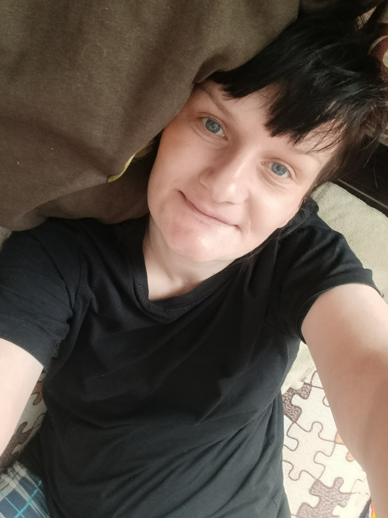 A light-skinned person with bright blue eyes and short, dark hair with bangs is captured in a high-angle selfie. They are lying down indoors, looking directly at the camera with a gentle, faint smile. The person is wearing a plain black, short-sleeved t-shirt. On the upper left, a brown or dark grey fabric, possibly a blanket or pillow, partially covers the person's left side. A small section of blue and white plaid fabric is visible on the lower left, likely part of their clothing or bedding. To the right of the person, a light-colored rug or mat with a distinctive jigsaw puzzle pattern (brown outlines on off-white pieces) is visible. Dark furniture or a headboard can be seen behind the person's head on the upper right. The setting suggests a casual indoor environment, possibly a bedroom, in Фоки, Russia. No specific time of day is discernible from the even indoor lighting, and there is no visible text in the image.