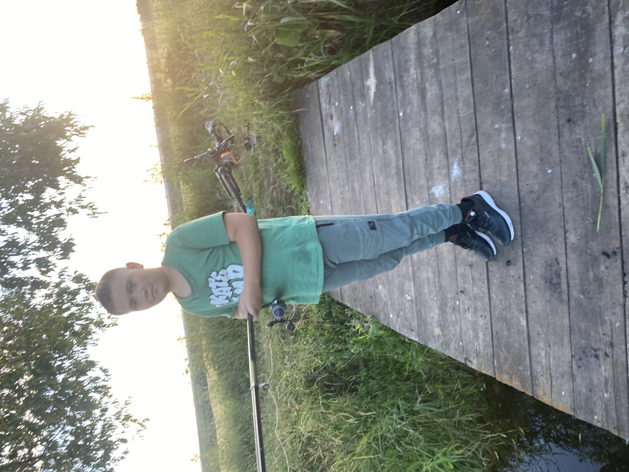 A young boy stands on a wooden pier next to a body of water, holding a fishing rod. He is wearing a green t-shirt and grey pants, with black sneakers. The pier is made of weathered wooden planks. To the right of the boy, a bicycle lies on the grass. The background is filled with tall green grass and foliage, and the setting appears to be a rural area. The sky is bright and overcast, suggesting it is daytime. The overall scene is natural and peaceful, with a focus on outdoor recreation. The location is the village of Afanasyevo, Russia.