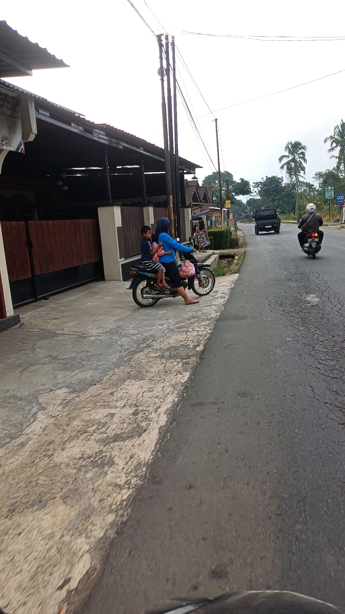 An overcast day in Tengahan, Indonesia, shows a mother and child riding a motorcycle along a paved road. The mother, wearing a blue headscarf and tunic, is driving the motorcycle, while a young boy, wearing a blue shirt and striped shorts, sits behind her. They are both wearing sandals. The motorcycle has a pink bag hanging from the front. They are on the left side of the road, next to a concrete sidewalk. On the sidewalk, there are a few small buildings with corrugated metal roofs and wooden fences. In the background, there are trees, palm trees, and more buildings. Another motorcycle with a person wearing a helmet is visible further down the road, along with a black pickup truck. The road surface is cracked and uneven.