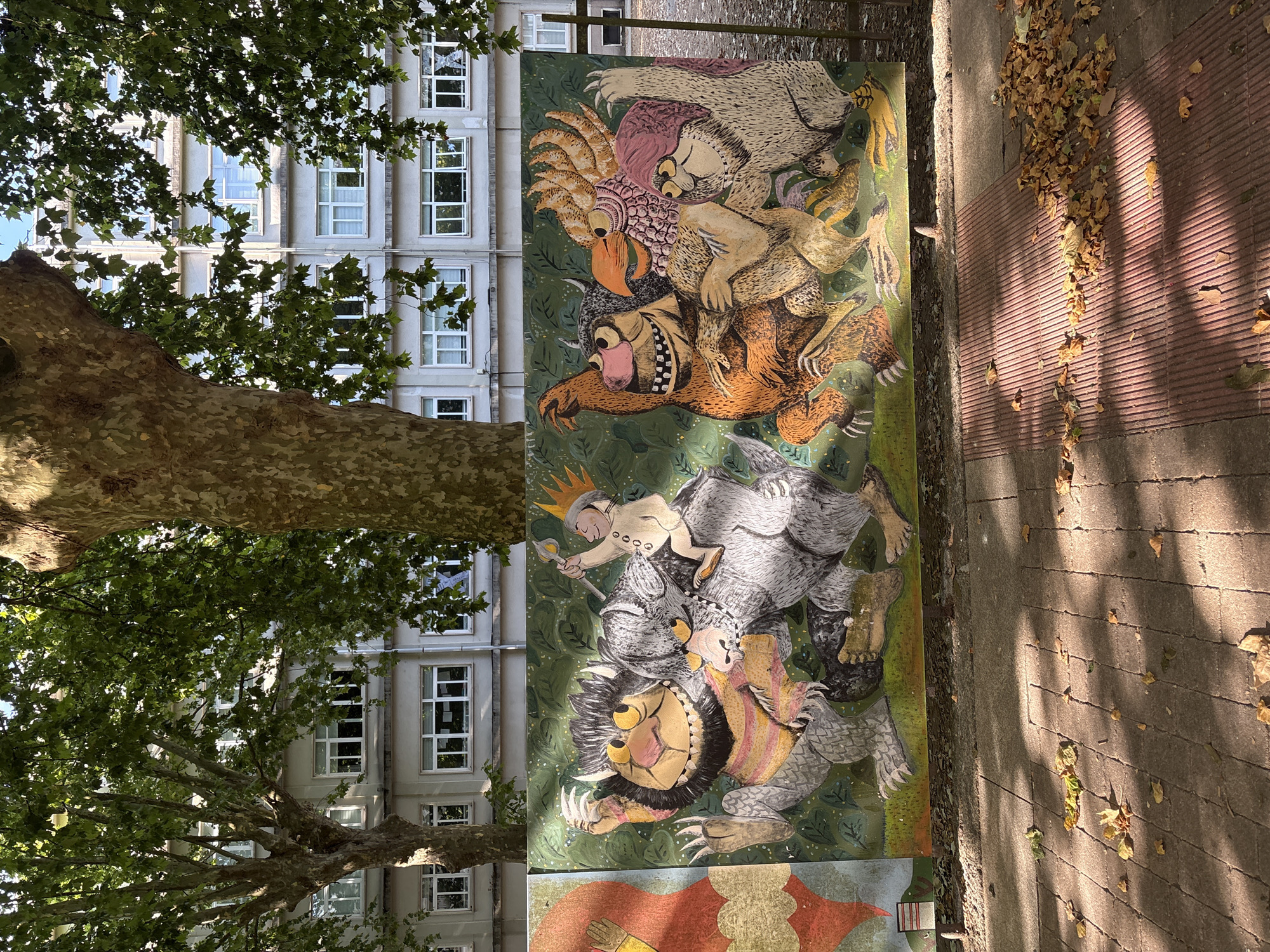 The image depicts a mural painted on a long, rectangular panel, installed outdoors, likely in a public space within the city of Pontevedra, Spain. The mural features characters from Maurice Sendak's "Where the Wild Things Are." On the left side of the mural, two large, furry, monster-like creatures are depicted, with one appearing to be riding on the back of the other. A small human-like figure with a crown is seated on the neck of the creature in the back, holding a stick with a flame on top. The creatures have distinct features, including large eyes, sharp teeth, and fur. To the right of these characters, a group of three more stylized, beast-like figures are shown, interacting with each other. These figures are also colorful and have unique appearances, with beaks, horns, and varying fur textures. The background of the mural is a dark green, with stylized leaf patterns.
The mural is positioned in front of a light-colored, multi-story building with numerous windows. In the foreground, a paved walkway, made of grey rectangular stones and some reddish ridged paving, is visible. Fallen autumn leaves are scattered across the paved area, suggesting a time of year when trees shed their foliage. Large trees with thick trunks and dense green foliage are situated to the left and right of the building and mural, casting shadows on the ground. The overall impression is an outdoor setting, possibly a park or a street lined with trees and a building. The mural's artistic style is illustrative, with bold lines and vibrant colors, evoking a sense of fantasy and childhood imagination.