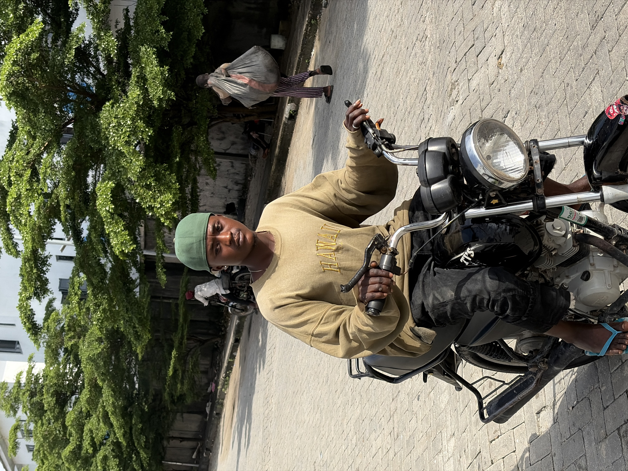 In Igboefon, Nigeria, a young Black male wearing a green cap and a tan sweatshirt with the word "HAWAII" embroidered in gold rides a black motorcycle. He is looking directly at the camera with a neutral expression. The motorcycle has a chrome headlight and handlebars. He is wearing black pants and light blue flip-flops. In the background, trees with lush green foliage line a paved street. Another person is visible in the background, walking away from the camera and carrying a large, grey bag over their shoulder. They are wearing striped pants and dark sandals. Further in the background, another person is on a motorcycle, also moving away from the camera. The scene appears to be a street in a city with buildings visible behind the trees. The pavement is made of interlocking bricks.