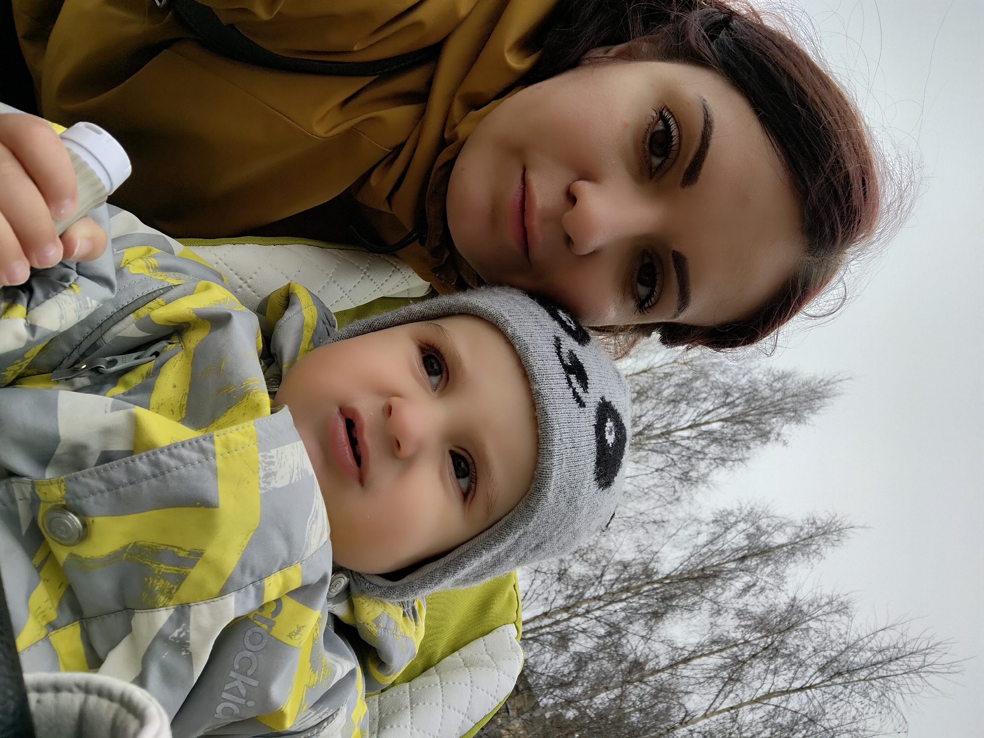 A woman and child are outdoors in what appears to be Markovskii, Russia, as suggested by the location request. The woman is positioned above the child, likely holding him or her. She has dark hair and eyes and wears a yellow jacket. The child wears a gray and yellow patterned snowsuit and a gray hat with a panda bear design. The child is also holding a bottle. The background features bare trees against a gray sky, indicating a winter or early spring season.