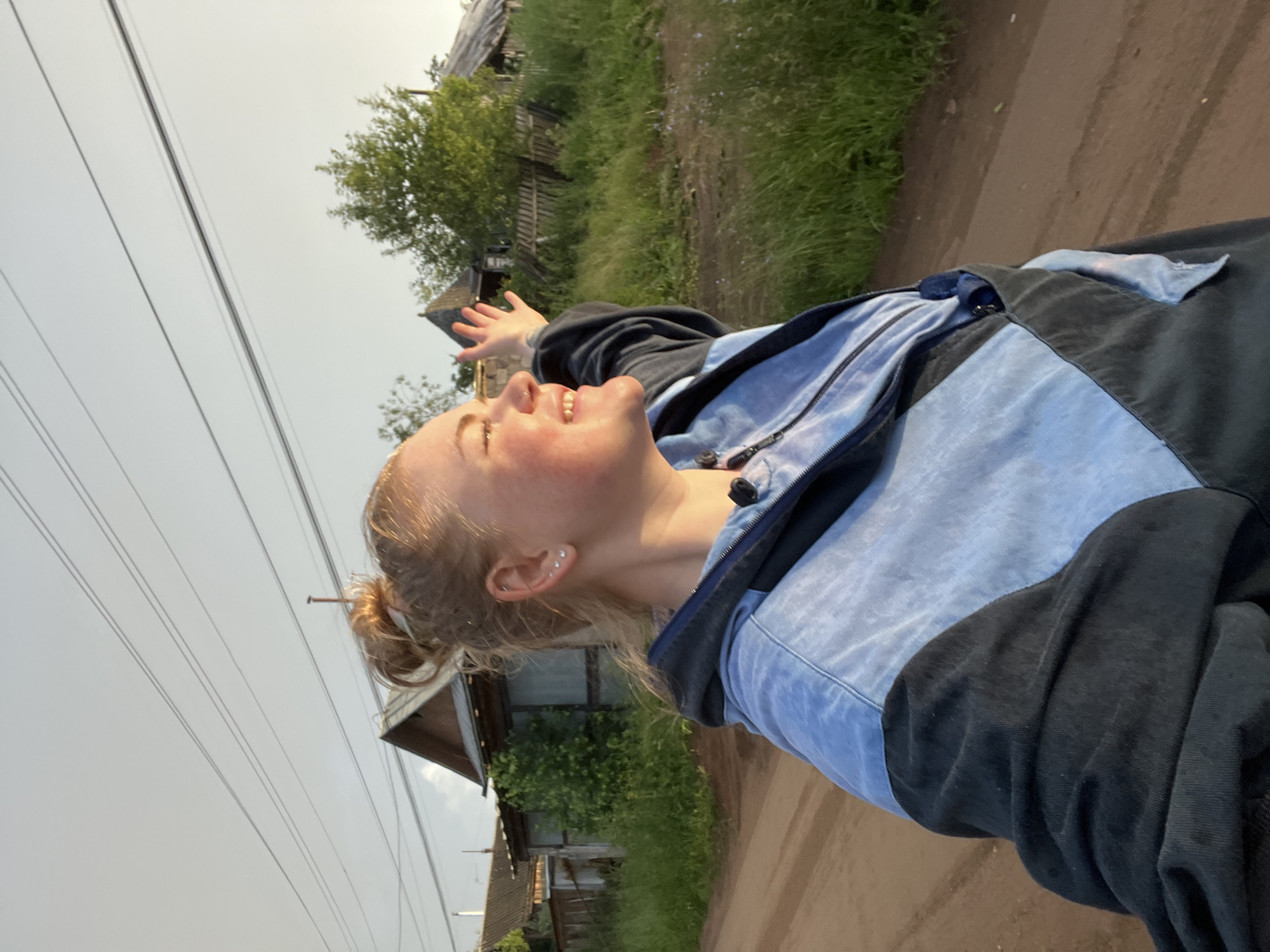 A young woman with blonde hair pulled back in a bun is captured in a close-up, low-angle shot from the chest up, outdoors in what appears to be a rural village. She is wearing a blue and black zip-up jacket. Her head is tilted back, and her eyes are closed with a radiant smile on her face, suggesting a moment of joy or exultation. Her left arm is raised with her palm open, as if embracing the environment.
The background shows a dirt road, lush green grass, and some rustic wooden houses, one of which has a weathered appearance. Trees are visible behind the houses, and a line of power lines stretches across the sky in the upper left quadrant of the image. The sky is overcast, indicating a potentially dull or rainy day, but the light on the woman's face suggests it might be late afternoon or early morning, with the sun trying to break through. The overall mood is one of simple happiness and freedom. The setting is identified as Olkhovka, Russia. There is no visible text in the image.