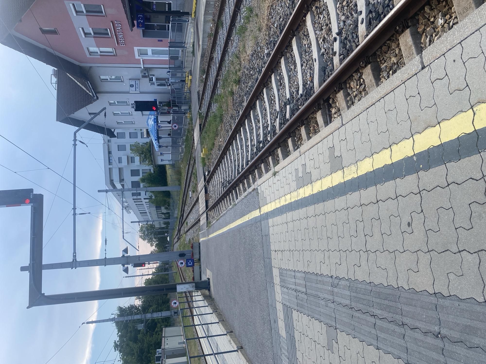 The image captures a view of the train station in Eisenstadt, Austria. The foreground features a train platform with patterned paving stones and a yellow safety line. Adjacent to the platform are railway tracks. In the background, a pink building with "EISENSTADT" signage is visible, along with other buildings and trees. Overhead electrical lines for the trains are prominent. A red train signal is illuminated. The sky appears overcast with some clouds. There are no visible people actively engaged in the scene. The overall environment depicts a functional train station setting.