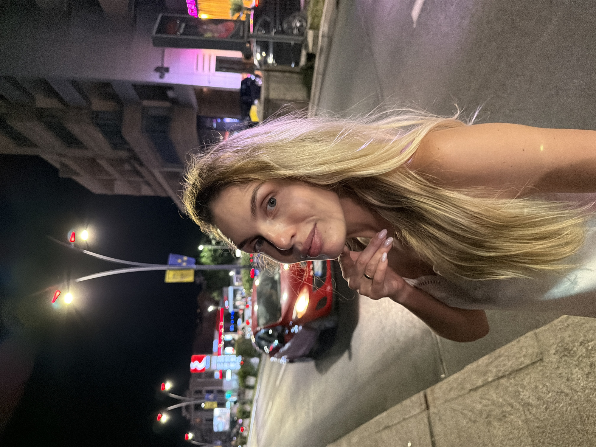 A woman with long blonde hair is captured in a low-light, nighttime outdoor scene, with a blurred urban background. She is positioned slightly off-center to the right, looking directly at the camera with a neutral expression. Her right hand is raised, with her fingers touching her chin and neck, and she wears a ring on her left ring finger. Her hair cascades down her shoulders, with some strands catching the ambient light. The environment behind her appears to be a city street with streetlights, buildings, and traffic. A red car is visible driving on the road, with its headlights on, and other vehicles and illuminated signs are present further in the background. The setting suggests a location in the city of Budva, Montenegro.