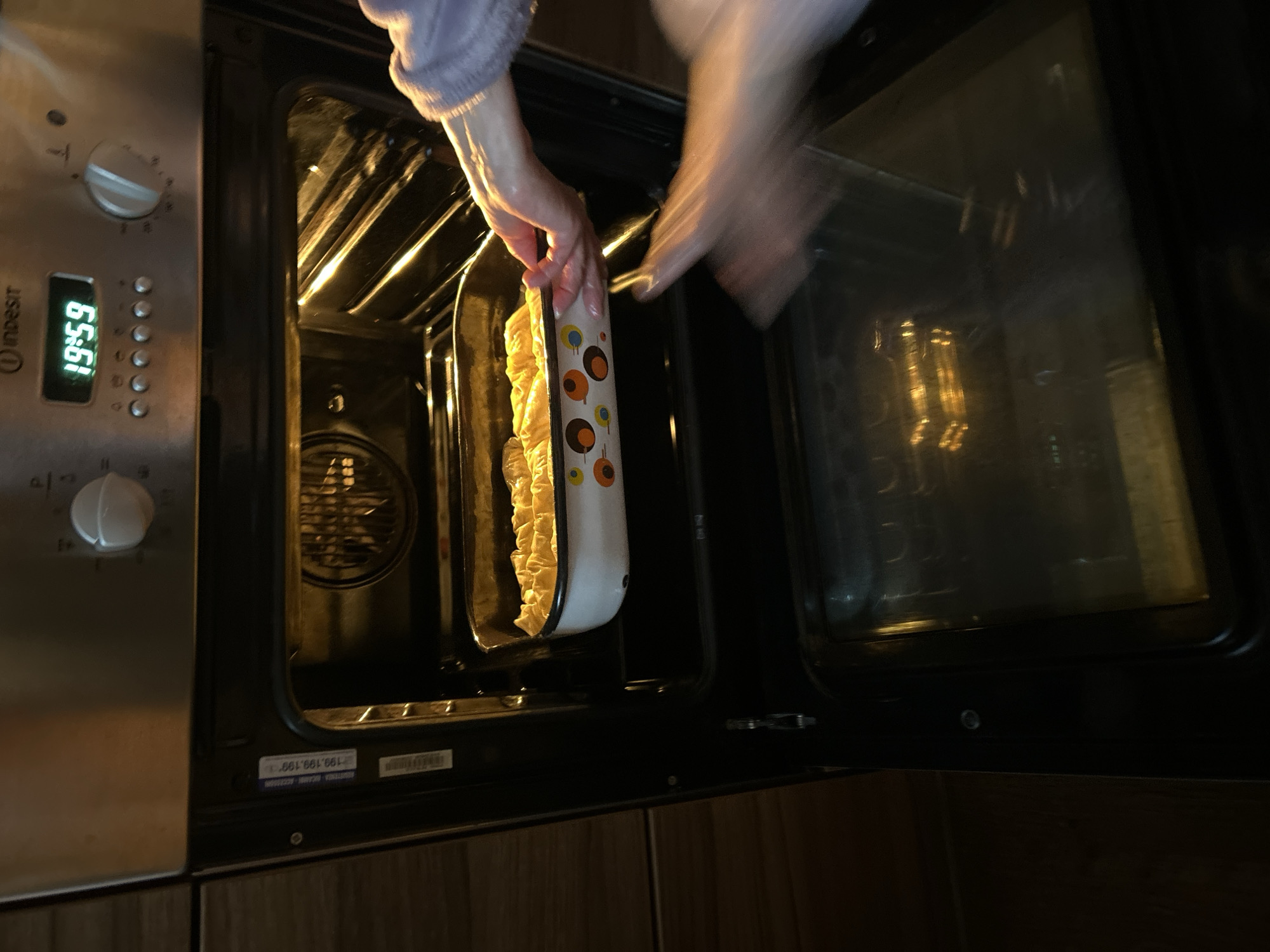 The image shows a person placing a baking dish into an open oven. The oven is stainless steel and black, with a digital clock displaying "19:59" visible on the left. The interior of the oven is lit, revealing racks. The baking dish is white with a patterned design. The environment includes a countertop with a wood-grain finish. The location is not discernible as Krumovo, Bulgaria.