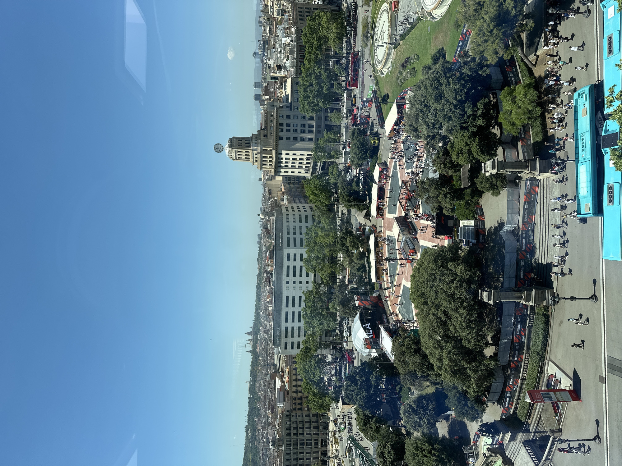 This is an aerial view of a public square in Barcelona, Spain, on a clear, sunny day. The square is bustling with activity. In the foreground, several bright turquoise buses are parked. In the middle ground, there is a large, open area with many people walking and standing. There are several temporary structures set up, including a stage with signage that reads "LEA." Surrounding the open area are numerous trees, providing shade. To the right of the square, a circular fountain with water features is visible, surrounded by a green lawn and more trees. In the background, various buildings of different architectural styles are visible, including a prominent building with a clock tower. The cityscape extends to a distant horizon with more buildings and hills. The overall scene depicts a vibrant urban environment with public events and a significant number of pedestrians.